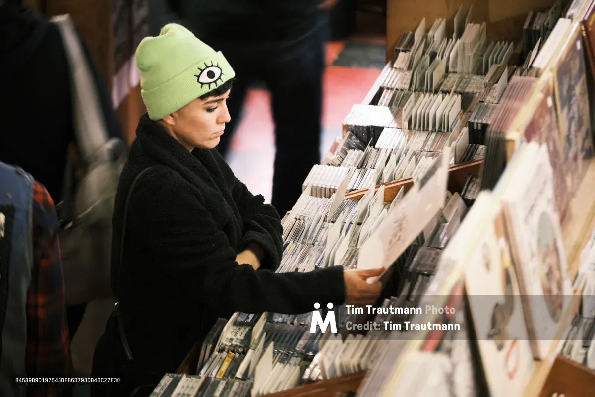 A young woman in a distinctive lime green beanie adorned with an eye symbol carefully examines vinyl records at Music Millennium during Record Store Day in Portland's Kerns neighborhood. The warm, amber lighting creates an intimate atmosphere as she browses through rows of meticulously organized albums, her black coat creating a stark contrast against the pale record spines. The scene captures the timeless ritual of record hunting on this celebrated day when music enthusiasts seek out special releases and support independent record stores.