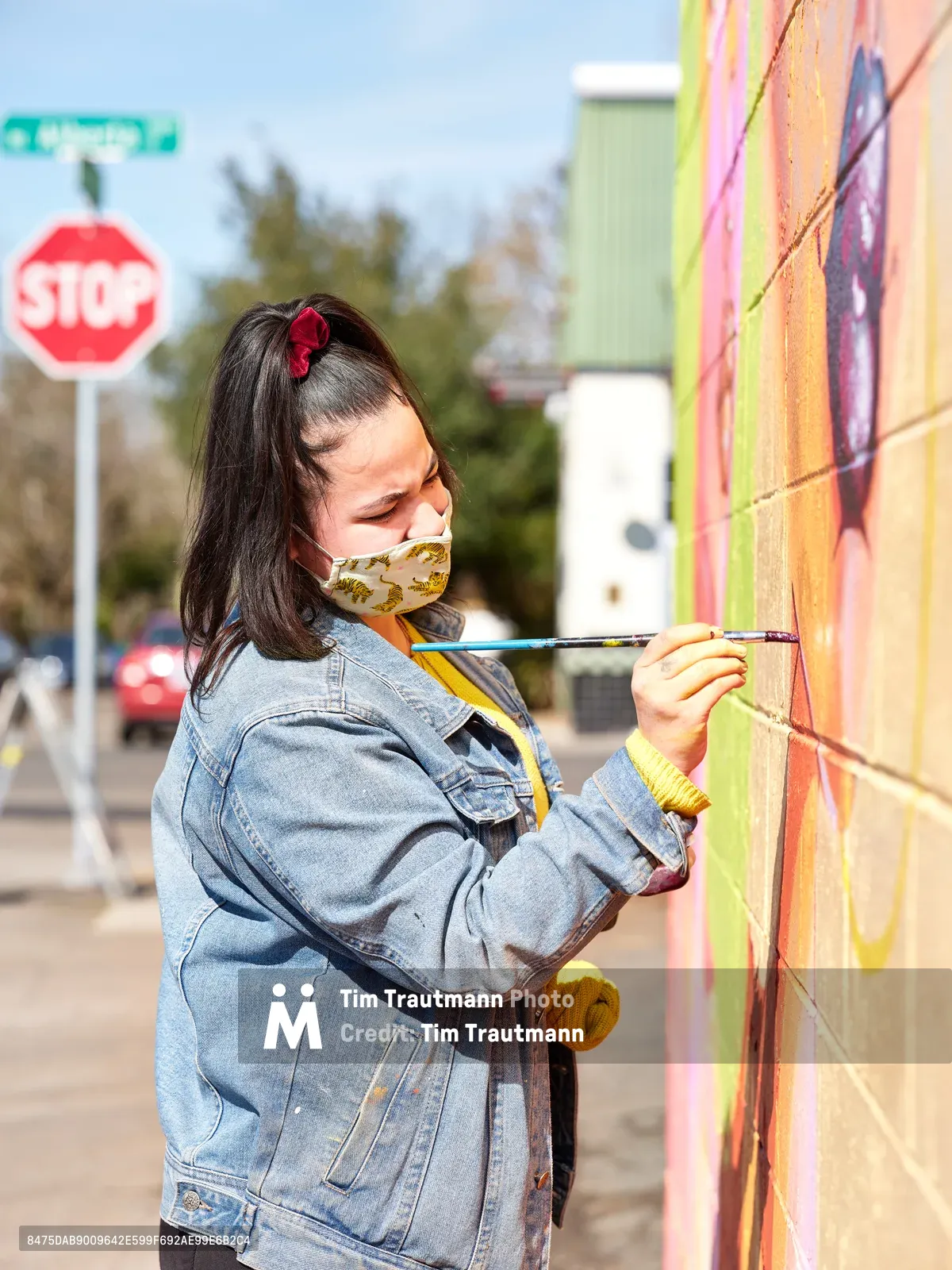 Multidisciplinary artist Vilatose carefully applies brushstrokes to her mural 'Jardín Cubana,' which depicts three female figures, on Alberta Street in Portland's bustling arts district. Wearing a denim jacket and floral face mask, she works with focused concentration against the warm afternoon light, her red hair accessory catching the sun. The urban intersection behind her, marked by a stop sign and residential buildings, frames this moment of creative expression in one of Portland's most vibrant cultural neighborhoods.
