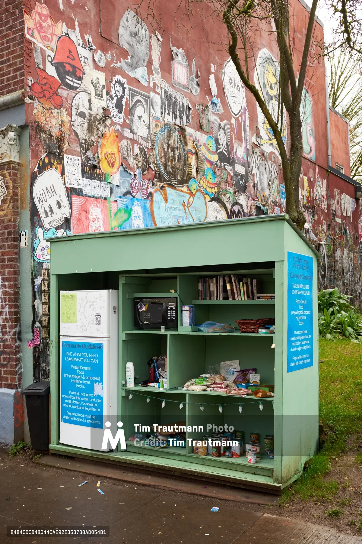 A sage green community refrigerator stands against a vibrant graffiti-covered brick wall on Alberta Street in Northeast Portland. The open pantry displays shelves stocked with donated food items, hygiene supplies, and reading materials, embodying grassroots mutual aid efforts. Blue informational signs explain the "take what you need, leave what you can" philosophy, while a leafless tree frames the scene in overcast daylight. The weathered urban setting contrasts with the organized generosity within the makeshift food bank.