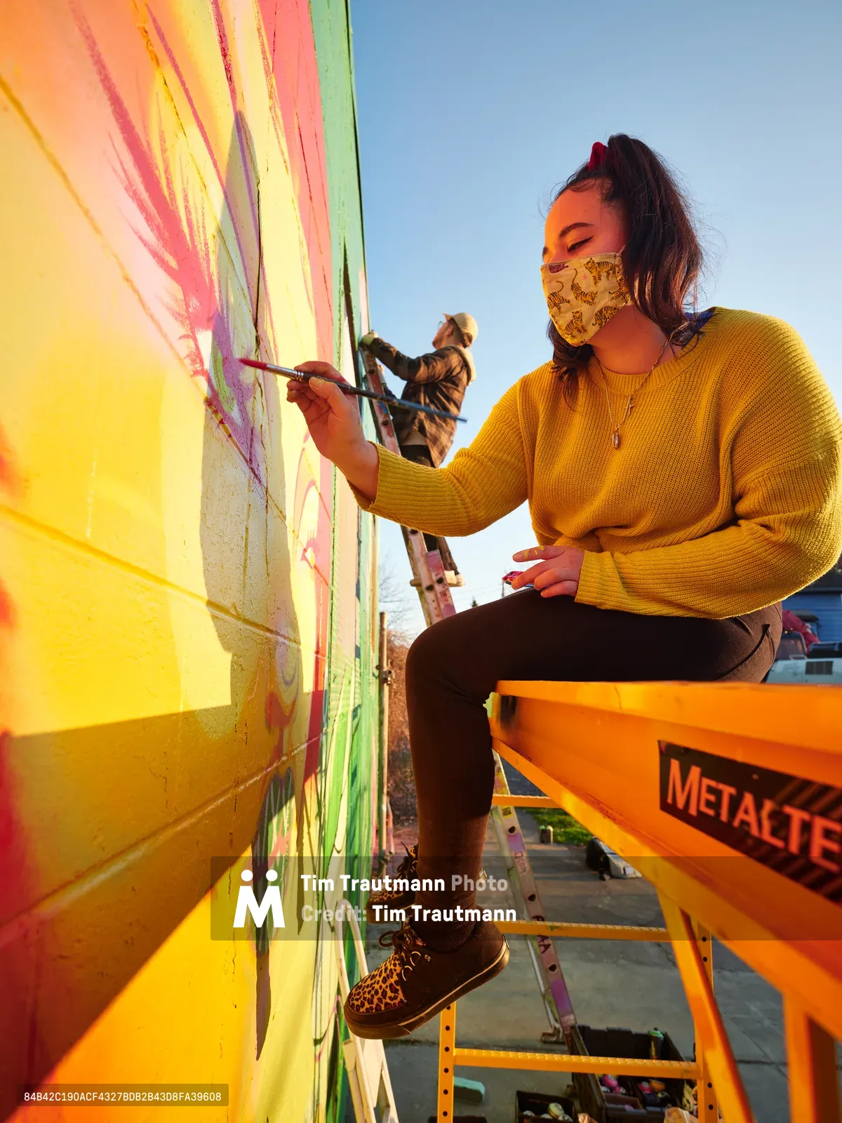 Multidisciplinary artist Vilatose applies vibrant brushstrokes to 'Jardín Cubana,' a mural depicting three female figures, while perched on an orange Metaltech scissor lift against the warm golden hour sky. The scene captures creative collaboration as another artist works in the background on the concrete wall. Wearing a patterned face mask and mustard yellow sweater, the artist embodies the resilient spirit of Portland's Alberta Arts District, where public art transforms urban landscapes into canvases of community expression.