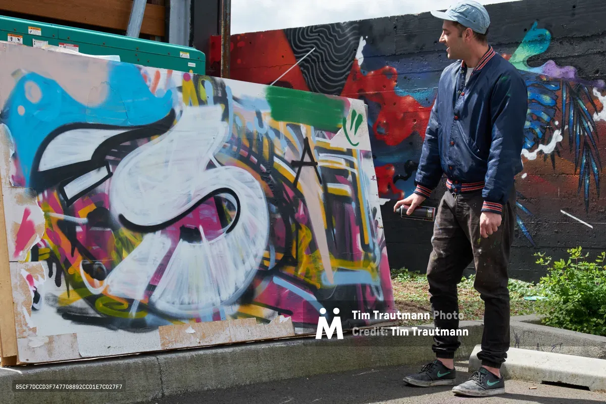 Galen Malcolm demonstrates spray painting techniques during a Portland Street Art Alliance tour at the Taylor Electric Blocks in southeast Portland's Hosford-Abernethy neighborhood. The artist, wearing a navy bomber jacket and gray cap, works on a vibrant piece featuring bold letterforms in blue, white, and pink against a colorful mural backdrop. Afternoon light illuminates the industrial setting where creative expression transforms urban walls into dynamic canvases.