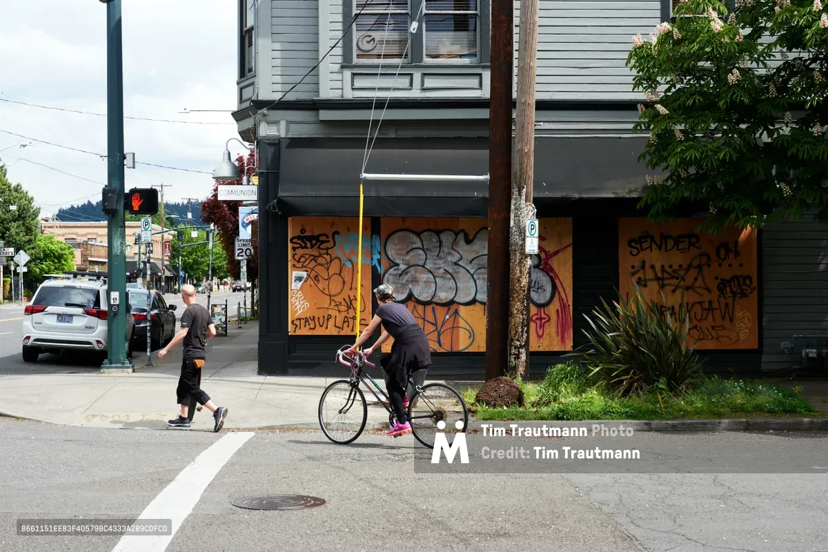 A cyclist in bright pink shoes pedals past a row of plywood-covered storefronts on Southeast Hawthorne Boulevard in Portland's Richmond neighborhood. Colorful graffiti tags sprawl across the orange and brown boards protecting the vacant businesses, while a pedestrian walks along the sidewalk under overcast skies. The scene captures the intersection of urban resilience and economic disruption in this historically vibrant commercial district.
