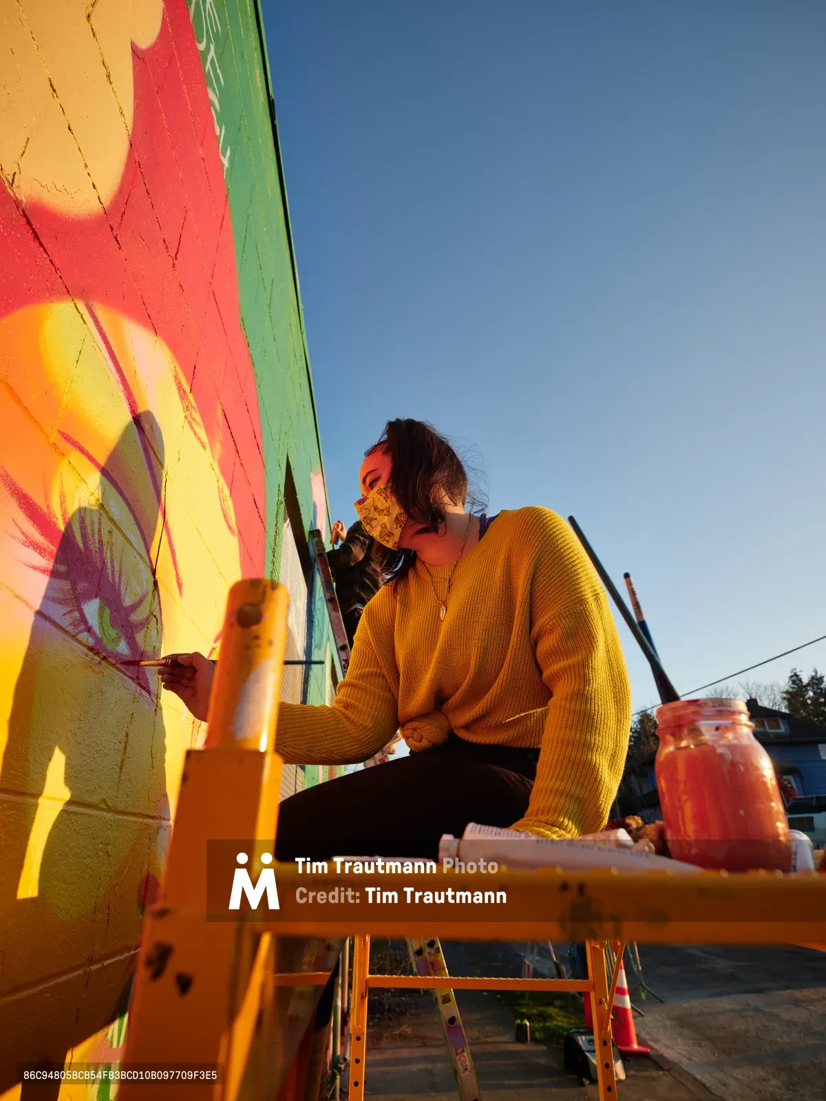 Multidisciplinary artist Villatose works from an orange scissor lift against a cloudless blue sky, painting the bold mural 'Jardín Cubana' featuring three female figures in tropical coral and yellow hues on Northeast Alberta Street. The masked artist, wearing a golden yellow knit sweater, concentrates on their work while surrounded by paintbrushes and materials in the heart of Portland's Alberta Arts District. The golden hour light bathes the scene in warm tones, highlighting the contrast between the vivid wall art and the serene evening atmosphere.