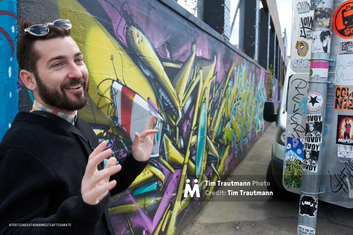 Tomás Valladares, a passionate guide from the Portland Street Art Alliance, gestures enthusiastically while explaining the vibrant graffiti mural behind him in the Taylor Electric Blocks. His animated expression and pointing hand draw viewers into the story of Portland's street art scene, while colorful wildstyle lettering in yellows and purples creates a dynamic backdrop. A utility pole covered in eclectic stickers stands sentinel beside the concrete walkway, testament to the grassroots creativity that defines Southeast Portland's Hosford-Abernethy neighborhood.