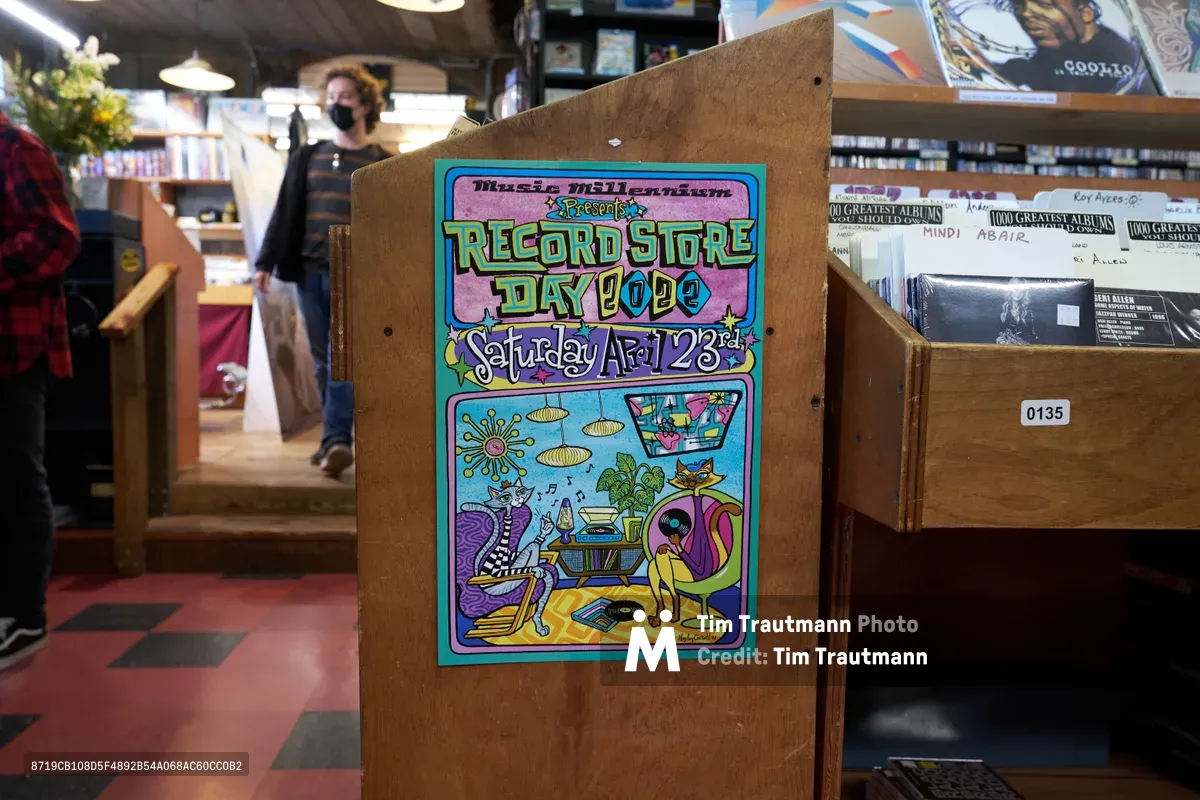 A vibrant hand-illustrated poster advertising Record Store Day 2022 stands prominently on a wooden easel inside Music Millennium, Portland's iconic independent record shop on East Burnside. The colorful artwork features whimsical characters in a psychedelic living room scene with floating UFOs and musical elements, embodying the creative spirit of vinyl culture. Behind the poster, the warm glow of pendant lighting illuminates rows of vinyl records and a masked customer browsing the collection, capturing the intimate atmosphere of this beloved neighborhood music sanctuary in the Kerns district.