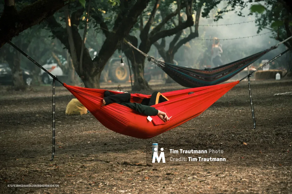 A person in black clothing reclines in a bright red hammock at the Orchard Vibration campout in Newberg, Oregon, with a canned drink resting on the ground below them. A second patterned hammock in blue and orange is visible in the background, suspended between large orchard trees. Hazy smoke drifts through the moody, dimly lit grove, with festival-goers, a tractor, string lights, and camping gear faintly visible among the trees.