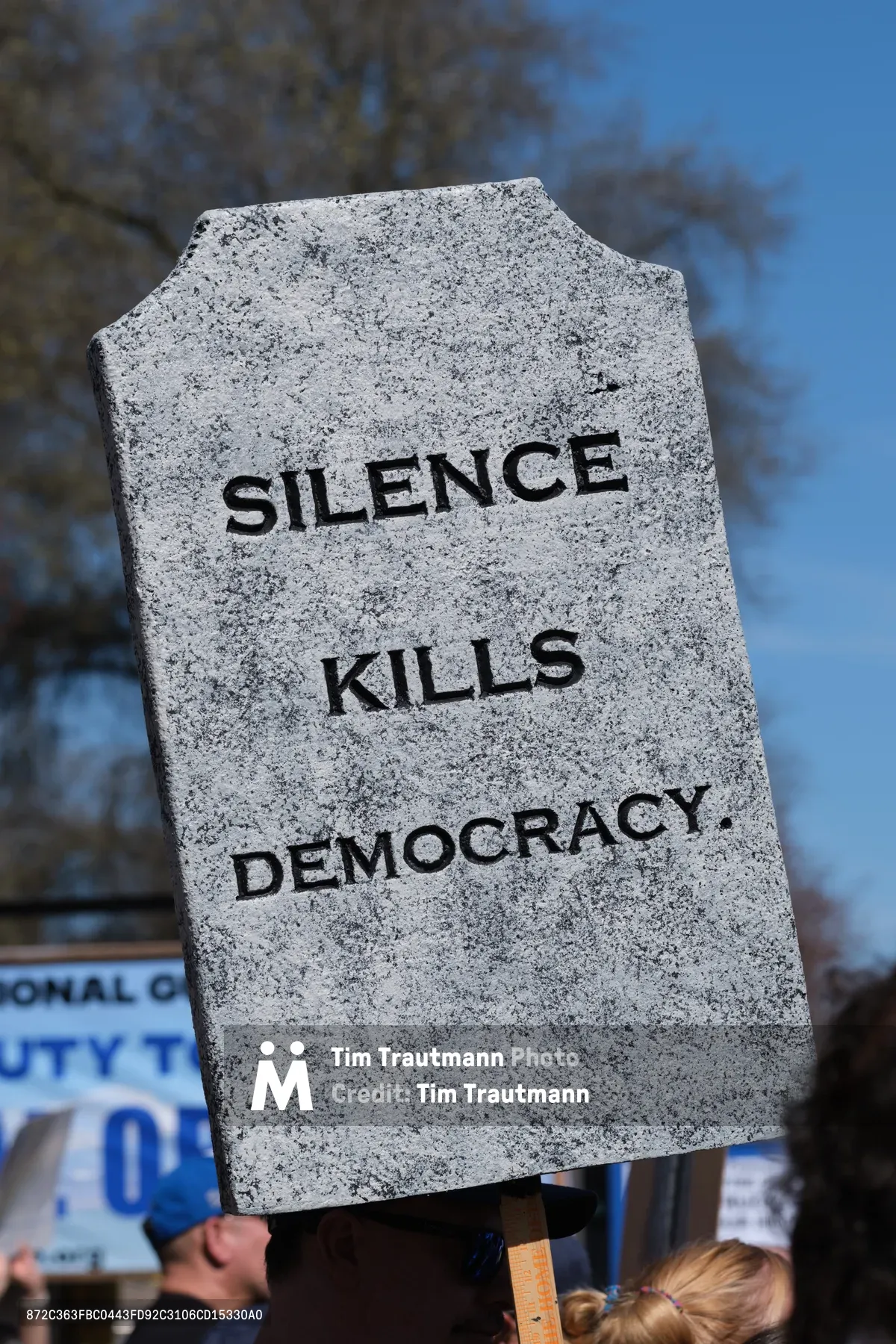 A weathered granite-textured protest sign shaped like a tombstone bears the stark message "SILENCE KILLS DEMOCRACY" in bold black lettering against Portland's azure winter sky. The foam board memorial, held aloft during the March 2026 No Kings demonstration in Old Town's historic Skidmore district, looms above glimpses of other protesters and campaign signage below. Bare tree branches frame the scene, creating a somber backdrop that amplifies the sign's grave warning about civic participation.
