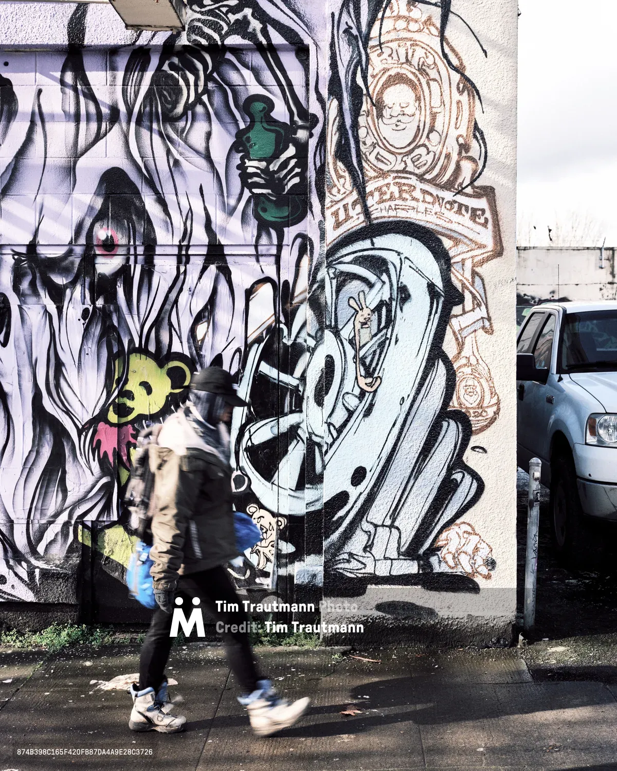 A motion-blurred pedestrian carrying a blue bag walks briskly along a wet sidewalk in Portland, Oregon, past a large-scale graffiti mural by artist Uter. The mural spans two building surfaces and features bold black-and-white surrealist imagery including a hooded figure, a cartoon bear character in yellow and pink, a stylized letter form with a rabbit inside, and circular emblems with illustrated faces. A parked white truck is partially visible on the right.