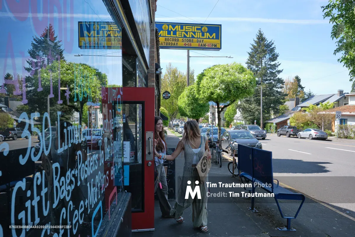 Two young women browse vintage records outside Music Millennium, Portland's iconic record store on East Burnside Street. The afternoon light casts gentle shadows across the sidewalk as one woman in a flowing dress examines vinyl while her companion watches from the doorway. The scene captures the enduring appeal of physical music media against the backdrop of Portland's eclectic Kerns neighborhood, where colorful storefronts and tree-lined streets create an authentic urban village atmosphere.