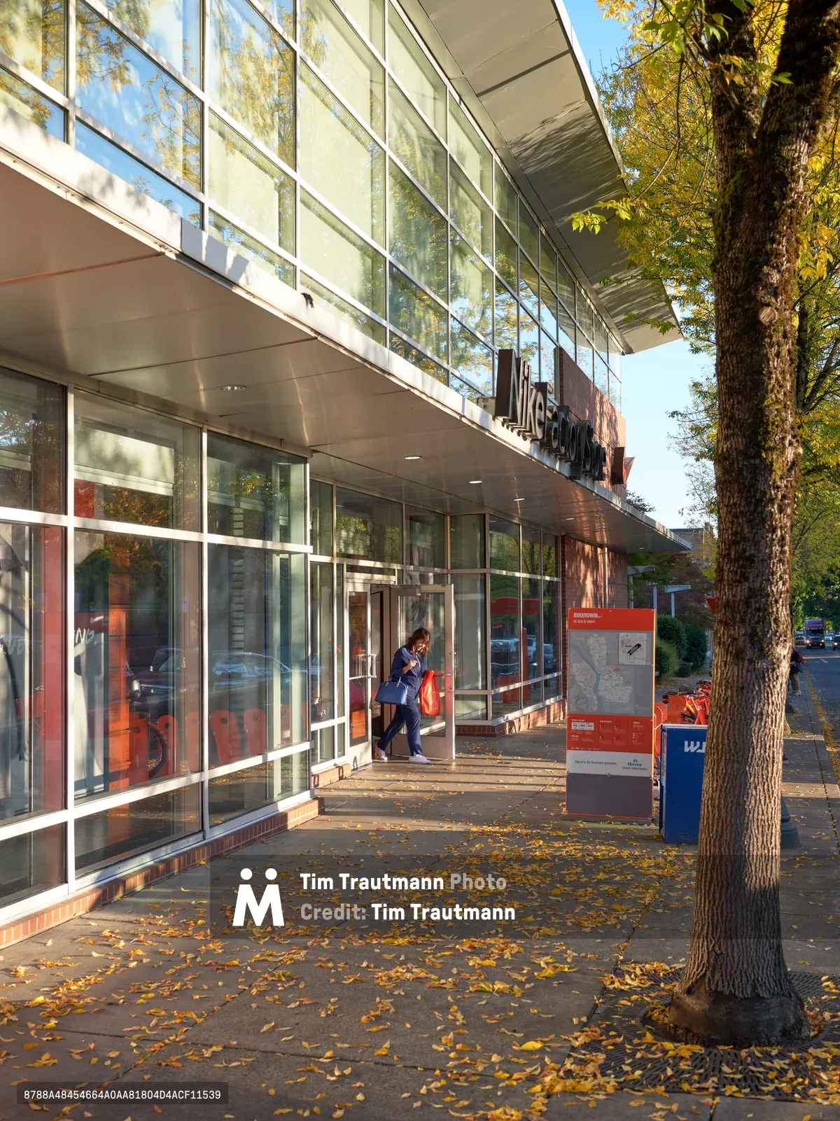 A lone shopper with a red bag walks through the modern glass-walled Nike Factory Store in Portland during golden hour autumn morning. Fallen yellow leaves carpet the concrete walkway beneath mature trees, while warm sunlight filters through the contemporary architecture's expansive windows. The scene captures the quiet rhythm of retail life as the store opens, with orange and blue Nike signage marking this retail destination.