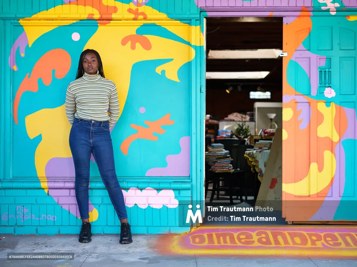 Naomi Likayi stands confidently beside her vibrant abstract mural on Northeast 15th Avenue in Portland's Alberta district. The young Black woman, dressed in a striped turtleneck and dark jeans, poses against the explosion of turquoise, yellow, coral, and lavender forms that transform the building's facade. The mural's organic shapes and playful palette create a striking contrast with the urban streetscape, while the open doorway reveals the interior of what appears to be a community space or shop.