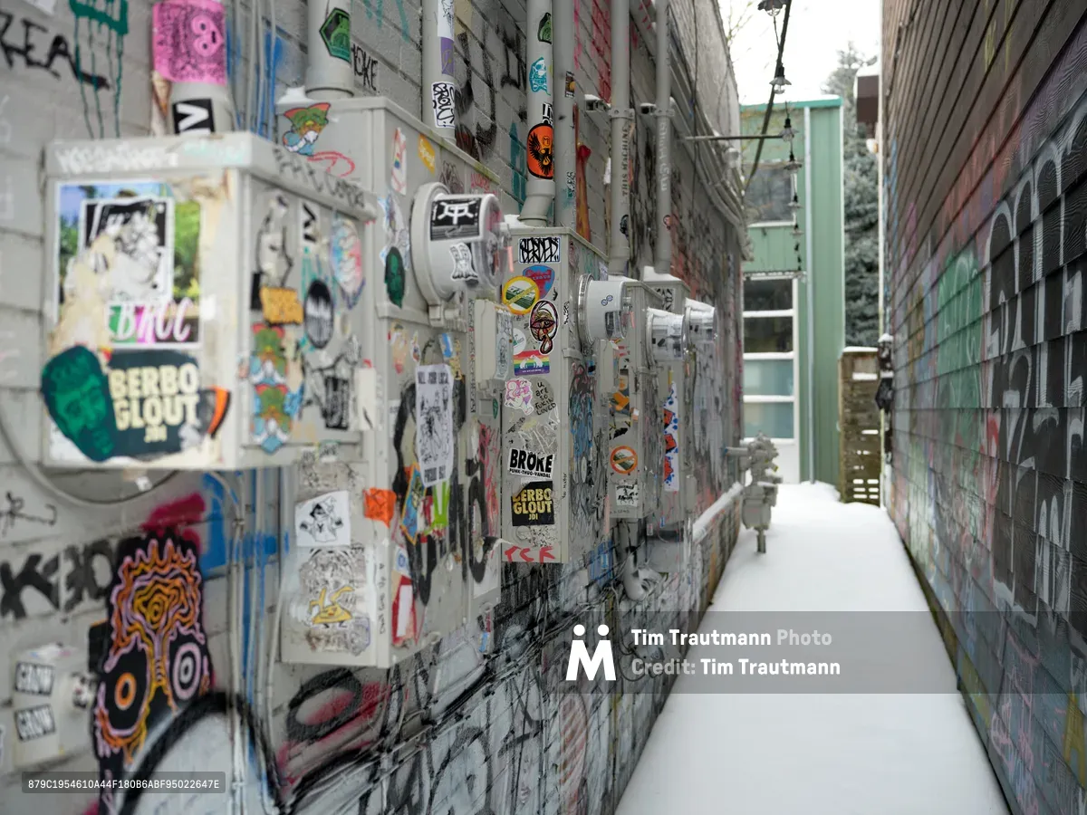 A narrow snow-dusted alleyway in Portland's Alberta Arts District transforms into an impromptu gallery where countless stickers and tags blanket electrical utility meters and concrete walls. The densely layered street art creates a vibrant patchwork of color against weathered gray surfaces, while fresh snow accumulates along the walkway edges. The perspective draws the eye down the corridor between buildings, where utility infrastructure becomes the canvas for an ever-evolving display of urban expression.