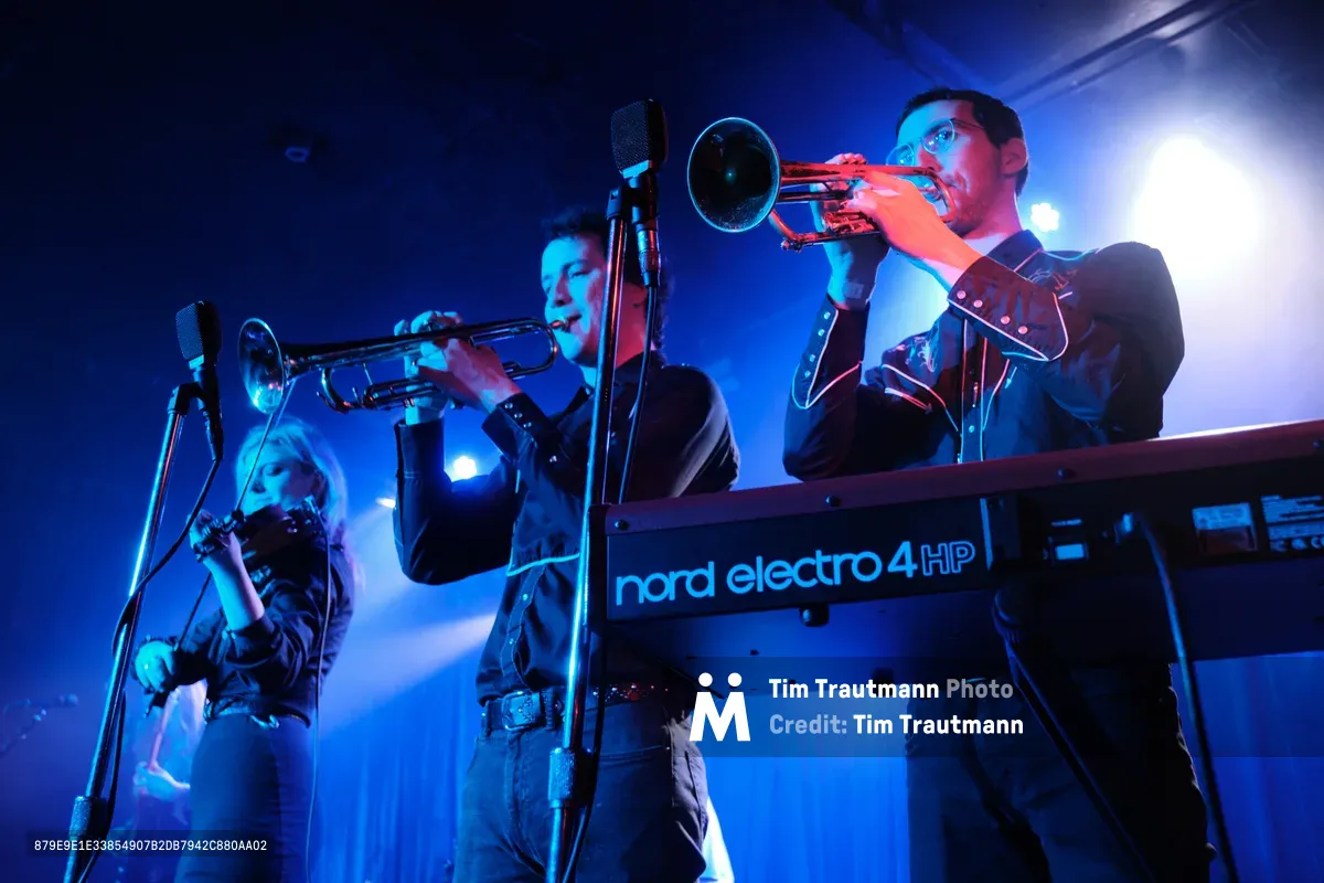 Two trumpet players from the band Roselit Bone perform in dramatic blue stage lighting at Dante's in Portland, Oregon during RX Fest 2019. The musicians, dressed in dark clothing, stand behind a Nord Electro 4 HP keyboard, their brass instruments gleaming under the moody concert illumination. Microphones capture their performance while intense backlighting creates an atmospheric haze, emphasizing the intimate venue setting of this underground music festival.