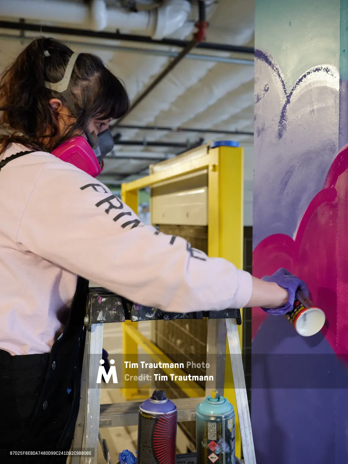 A focused artist in protective mask and gloves spray-paints vibrant purple and magenta forms onto a wall within the industrial confines of Clay Creative's parking garage in Southeast Portland. The scene captures the intersection of urban art and creative workspace, with aluminum ladders and spray cans arranged around the emerging mural. Warm artificial lighting illuminates the work in progress, creating an intimate documentation of street art creation in Portland's Hosford-Abernethy neighborhood.