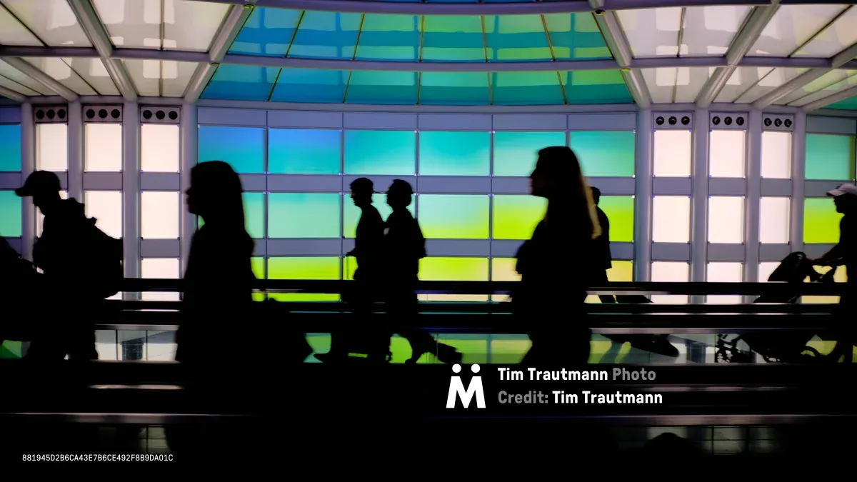 Travelers are silhouetted against the vivid backlit art installation "The Sky's the Limit" in the underground pedestrian tunnel connecting terminals at Chicago O'Hare International Airport. The curved ceiling and wall panels glow in shifting bands of turquoise, green, and yellow, creating a striking contrast against the dark figures of passing passengers. Their reflections shimmer on the polished floor below.