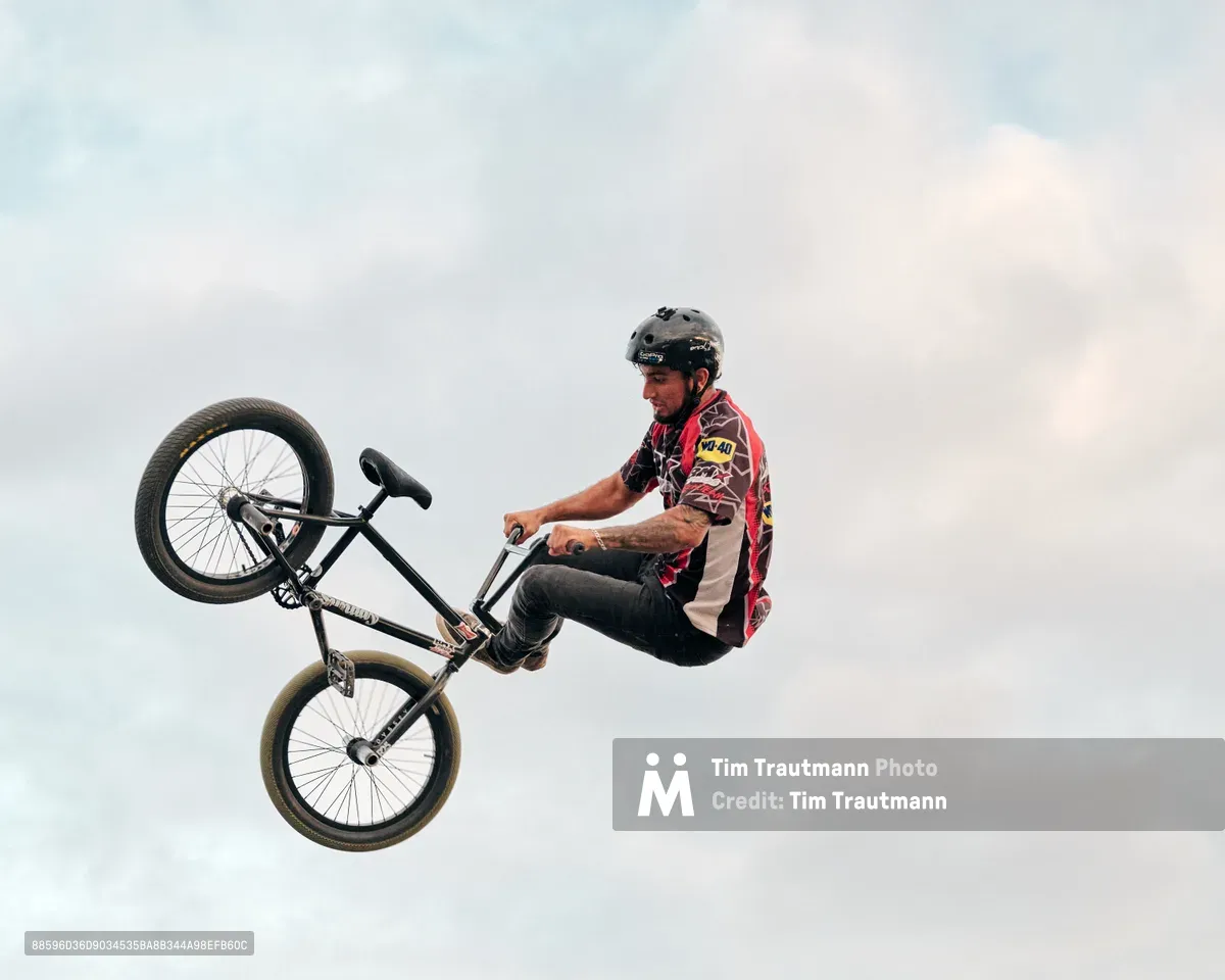 A BMX rider executes a dramatic aerial maneuver against the pale Washington sky at the Clark County Fair in Vancouver. The athlete, wearing a distinctive red racing jersey and protective helmet, grips his black bicycle while suspended mid-flight during what appears to be a freestyle demonstration. The overcast afternoon light creates a soft, ethereal backdrop that emphasizes the gravity-defying nature of the stunt, capturing a moment of athletic prowess and controlled risk at this beloved Pacific Northwest community gathering.