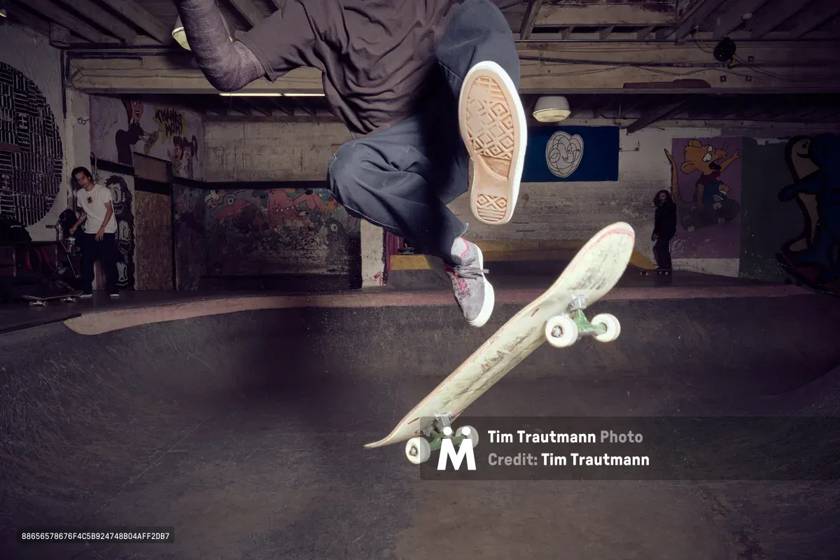 A skateboarder's legs and board are captured mid-air above the curved concrete of Commonwealth Skateboarding's bowl in Portland's Buckman neighborhood. The atmospheric lighting illuminates weathered sneaker soles and the deck's underside while graffiti-covered walls and fellow skaters create an authentic underground scene. The low-angle perspective emphasizes the height and technical precision of the aerial maneuver.