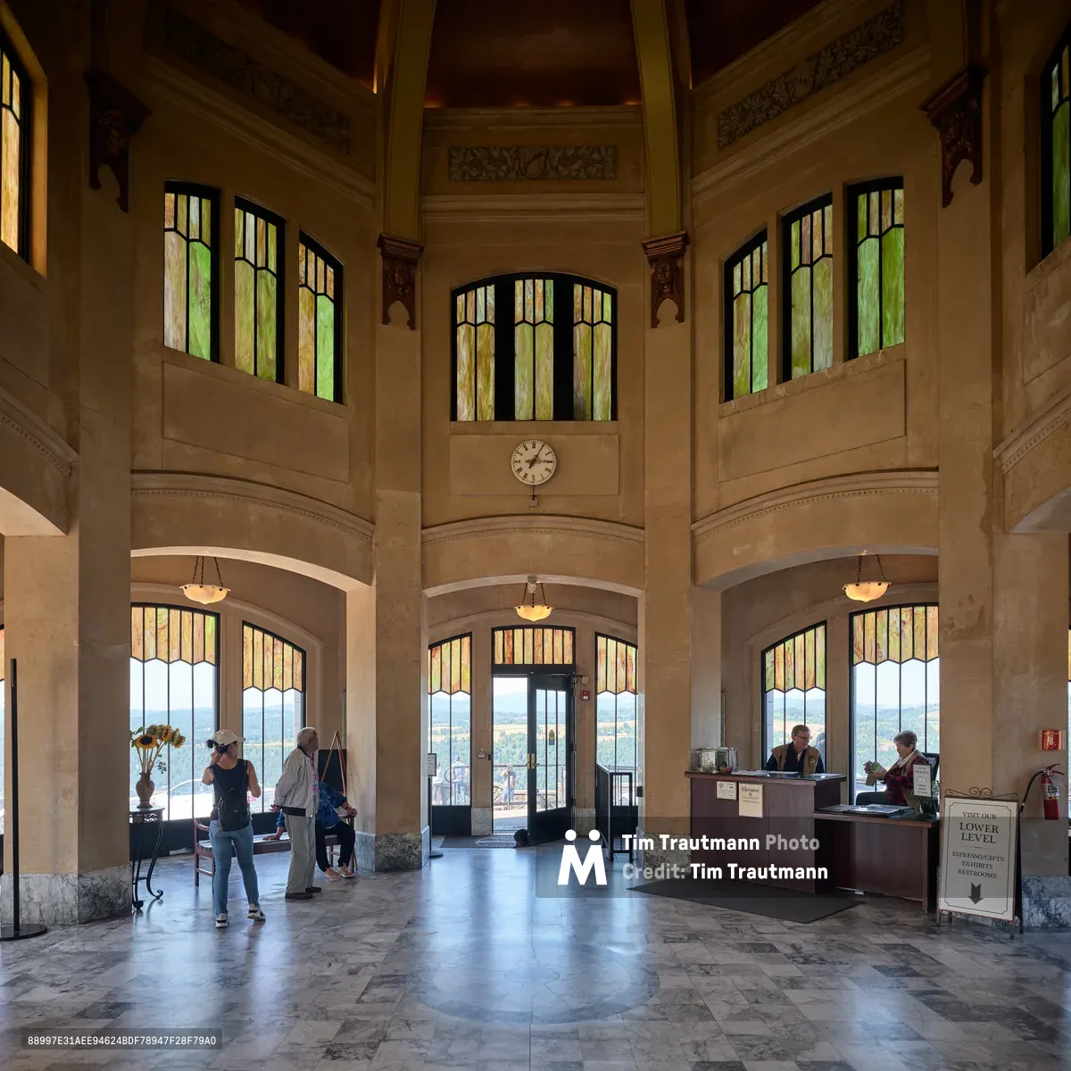 The soaring interior of Vista House reveals its 1918 architectural grandeur, with honey-toned stone walls rising to an octagonal crown adorned with stained glass windows. Visitors move quietly through the formal space, their footsteps echoing across polished marble floors as natural light filters through amber and green glass panels. An information desk anchors the ceremonial hall, while the central clock marks time beneath ornate carved details that speak to the structure's role as both scenic overlook and architectural monument.
