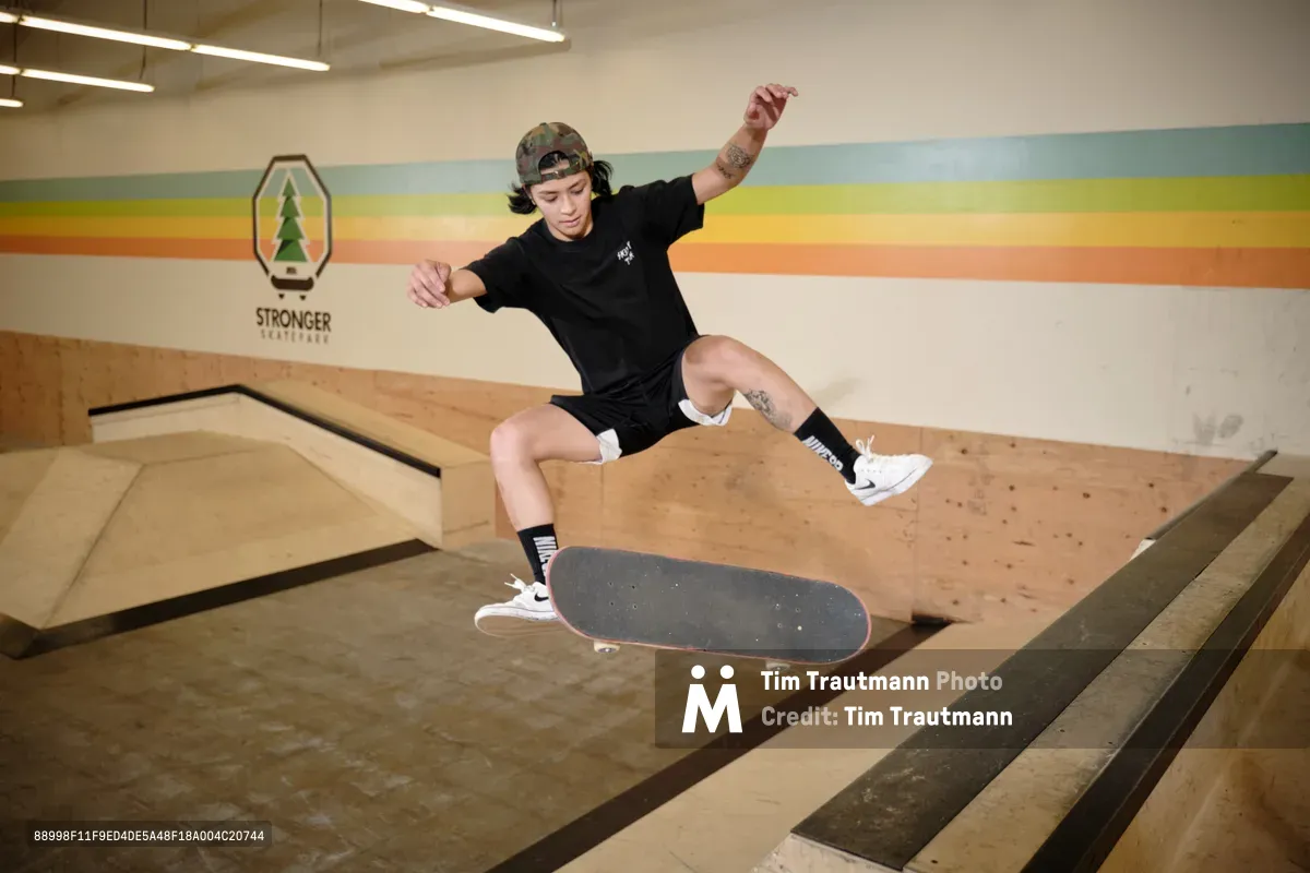 Professional skateboarder Christiana Means executes a dynamic aerial maneuver above the wooden bowl at Stronger Skatepark in Milwaukie, Oregon. Suspended in mid-flight with arms outstretched for balance, she demonstrates precise board control against the venue's distinctive rainbow-striped wall mural. The warm fluorescent lighting illuminates the polished plywood surfaces and creates an intimate atmosphere within this Pacific Northwest skateboarding sanctuary.