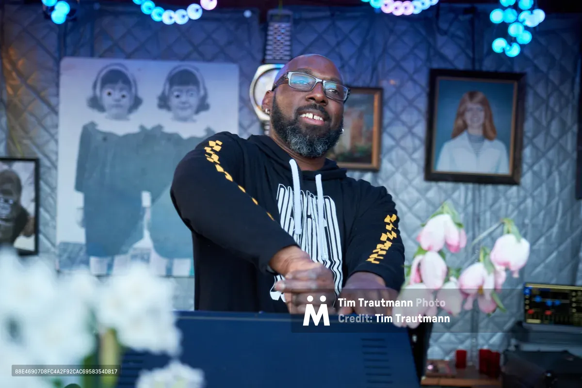 DJ Cee White, wearing glasses and a black graphic hoodie with yellow accents, performs with animated expression at the microphone during the Your Sunday Best season opener at White Owl Social Club in Portland. The intimate venue glows with blue-tinted lighting, creating atmospheric bokeh that dances across vintage portrait artwork adorning the diamond-patterned walls. Pink flowers in the foreground add a soft contrast to the moody blues and shadows of this quintessential Portland nightlife scene.