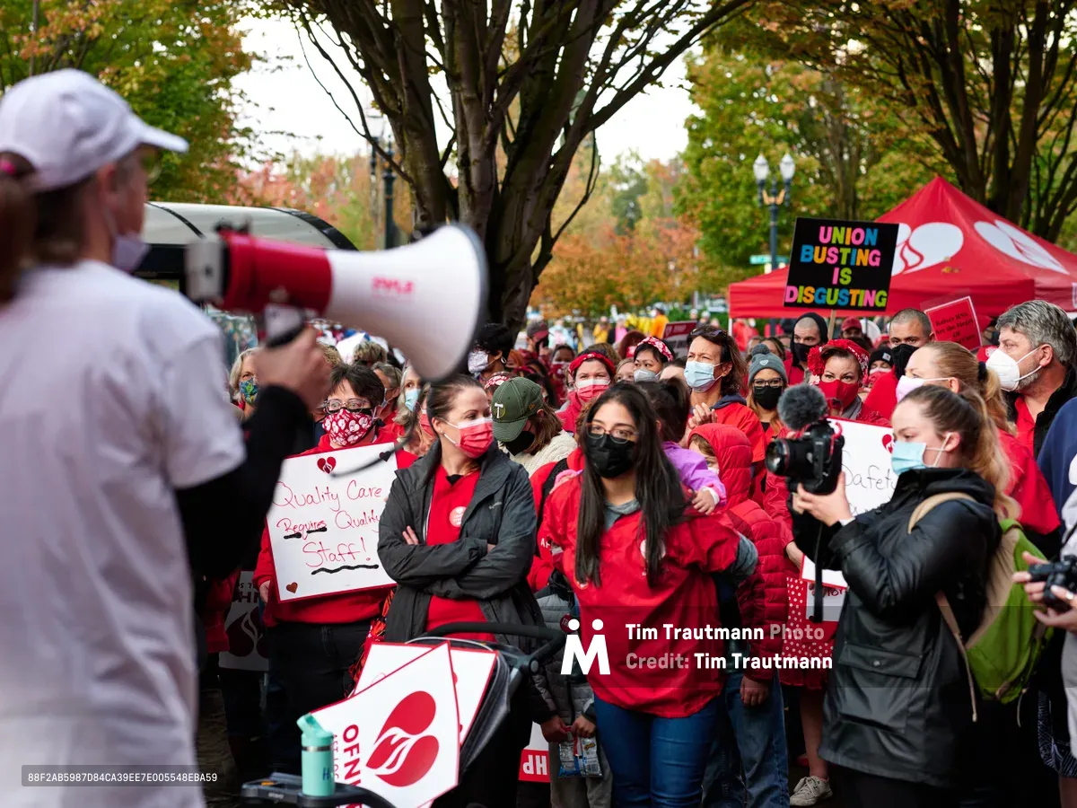 A sea of masked healthcare workers in crimson scrubs and winter jackets gather beneath autumn trees in Portland's Lloyd District, their unified stance punctuated by handmade signs demanding quality care and fair staffing. A speaker with a red and white megaphone commands attention from the foreground, while the crowd extends toward a vibrant tent displaying union solidarity messages. The overcast sky and golden fall foliage create a dramatic backdrop for this moment of collective action outside Kaiser Permanente's tower.