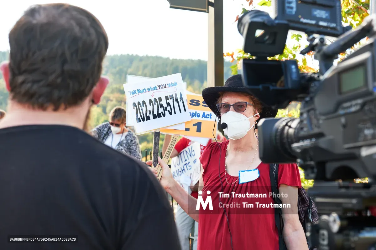 A determined climate activist in a red blouse and black hat addresses KOIN Channel 6 news cameras during a protest at Representative Kurt Schrader's Oregon City office. Behind her, fellow demonstrators hold signs including a phone number urging action, while autumn sunlight filters through the trees of McLoughlin Promenade. The scene captures the intersection of grassroots activism and media coverage, with the activist's white mask and focused demeanor conveying the urgency of climate advocacy in the Pacific Northwest.