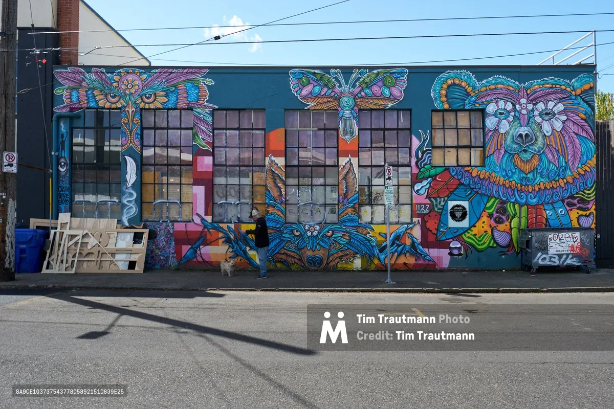 A magnificent large-scale mural transforms an industrial warehouse facade in Portland's Central Eastside district, featuring vibrant psychedelic animals rendered in intricate mandala patterns. The artwork showcases an owl with piercing eyes, a majestic bear, butterflies, and other creatures painted in electric blues, purples, oranges, and greens against the building's teal base. A lone pedestrian walks past the multi-windowed structure, providing human scale to this explosion of street art creativity that epitomizes the neighborhood's artistic renaissance.