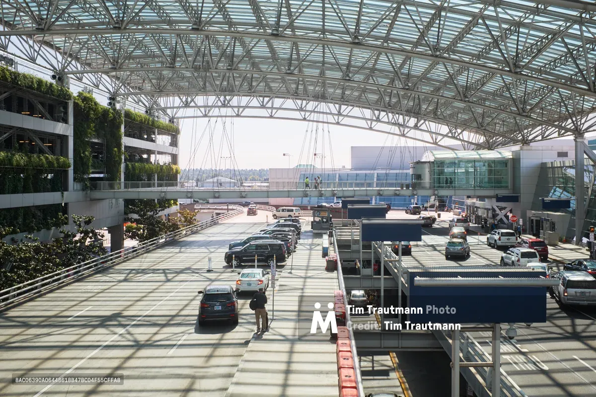 Beneath Portland International Airport's soaring latticed canopy, the departures driveway bustles with purposeful activity as travelers navigate the covered drop-off zone. The geometric steel framework creates dramatic shadows across the concrete surface while cars queue in organized lanes, their occupants preparing for journeys ahead. Green living walls on the terminal's left side soften the industrial architecture, while the distant mountains provide a serene backdrop to this choreographed transportation hub.
