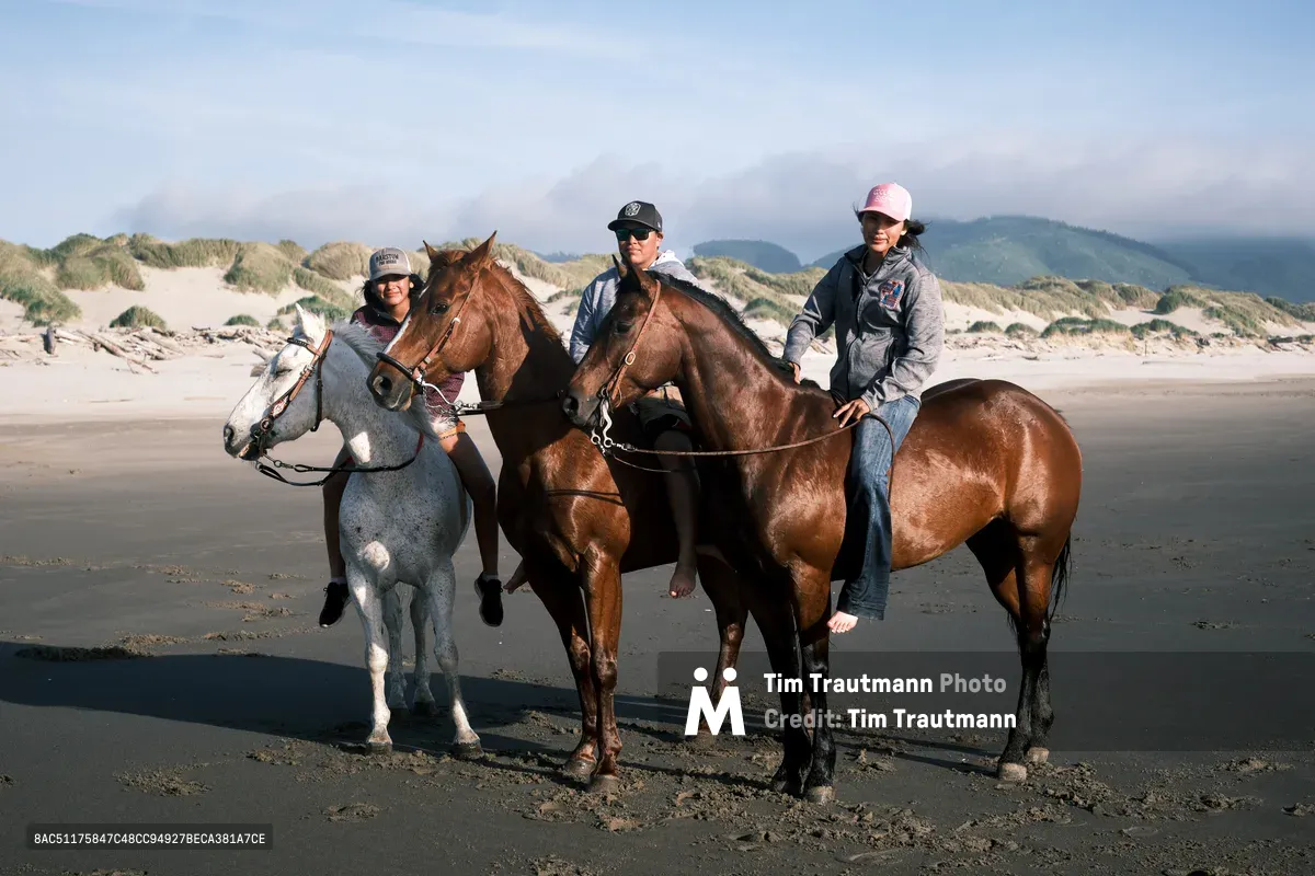 Three equestrians pause their beach ride along Oregon's dramatic coastline near Wheeler, their mounts - a dappled gray and two chestnuts - standing against the moody Pacific horizon. Rolling sand dunes frame the scene while distant coastal mountains emerge through soft maritime haze. The riders, casually dressed in baseball caps and hoodies, embody the relaxed spirit of Pacific Northwest beach culture, their horses' hooves having traced temporary paths across the tide-compacted sand.