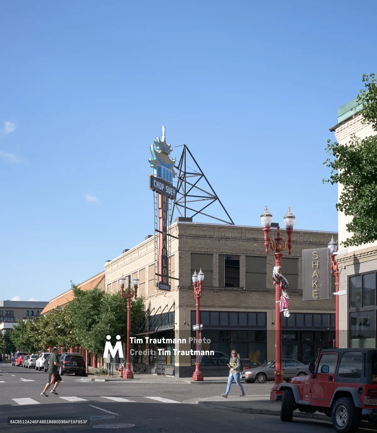 A pedestrian crosses the sun-drenched intersection in Portland's historic Chinatown district, where traditional pagoda-style signage crowns weathered brick buildings. The clear blue afternoon sky illuminates the distinctive red ornamental streetlamps and architectural details that define this cultural neighborhood. A red Jeep waits at the crosswalk as daily life unfolds along the tree-lined commercial strip, where vintage neon signs hint at the area's storied past.