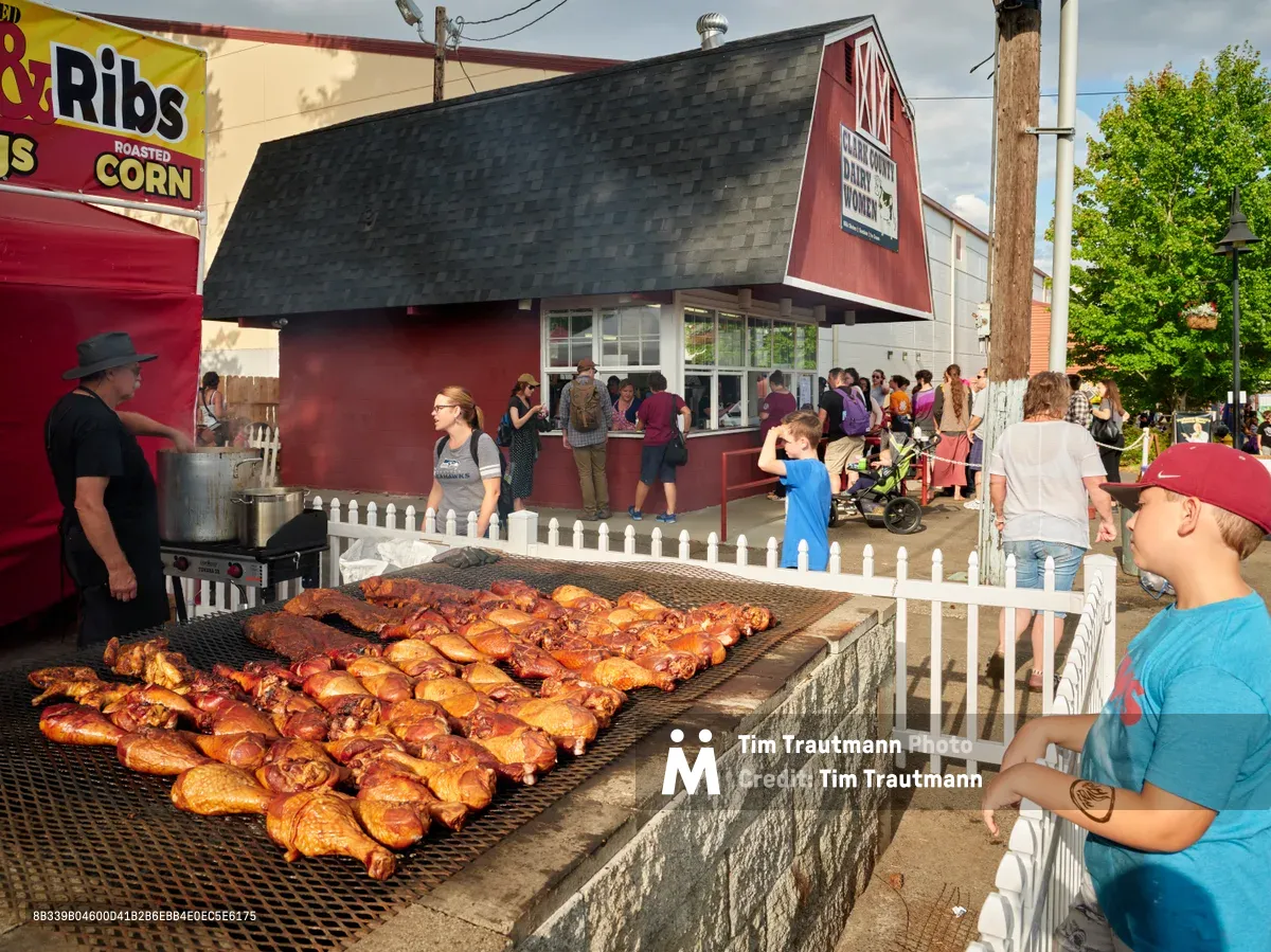 Golden-brown ribs and sausages sizzle on a large outdoor grill at the Clark County Fair in Vancouver, Washington, their caramelized surfaces glistening under the afternoon light. A pitmaster in black tends to the massive stone barbecue pit while fairgoers, including children and families, gather behind white picket fencing to watch the culinary spectacle. The scene captures the quintessential American county fair atmosphere, with red barn-style buildings and food vendor banners creating a festive backdrop amid the summer crowd.