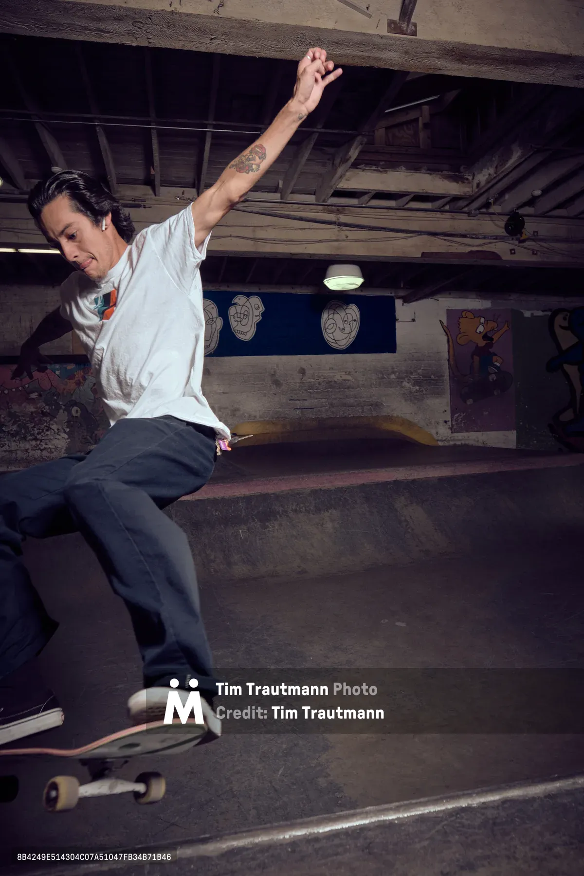A tattooed skateboarder launches into motion within the gritty confines of Commonwealth Skateboarding's basement venue in Portland's Buckman neighborhood. The raw industrial space, with its exposed wooden beams and graffitied walls, creates a moody backdrop as harsh overhead lighting cuts through the underground atmosphere. The skater's white long-sleeve shirt contrasts sharply against the weathered concrete and dark shadows, while cartoon murals peek through the dimly lit periphery.