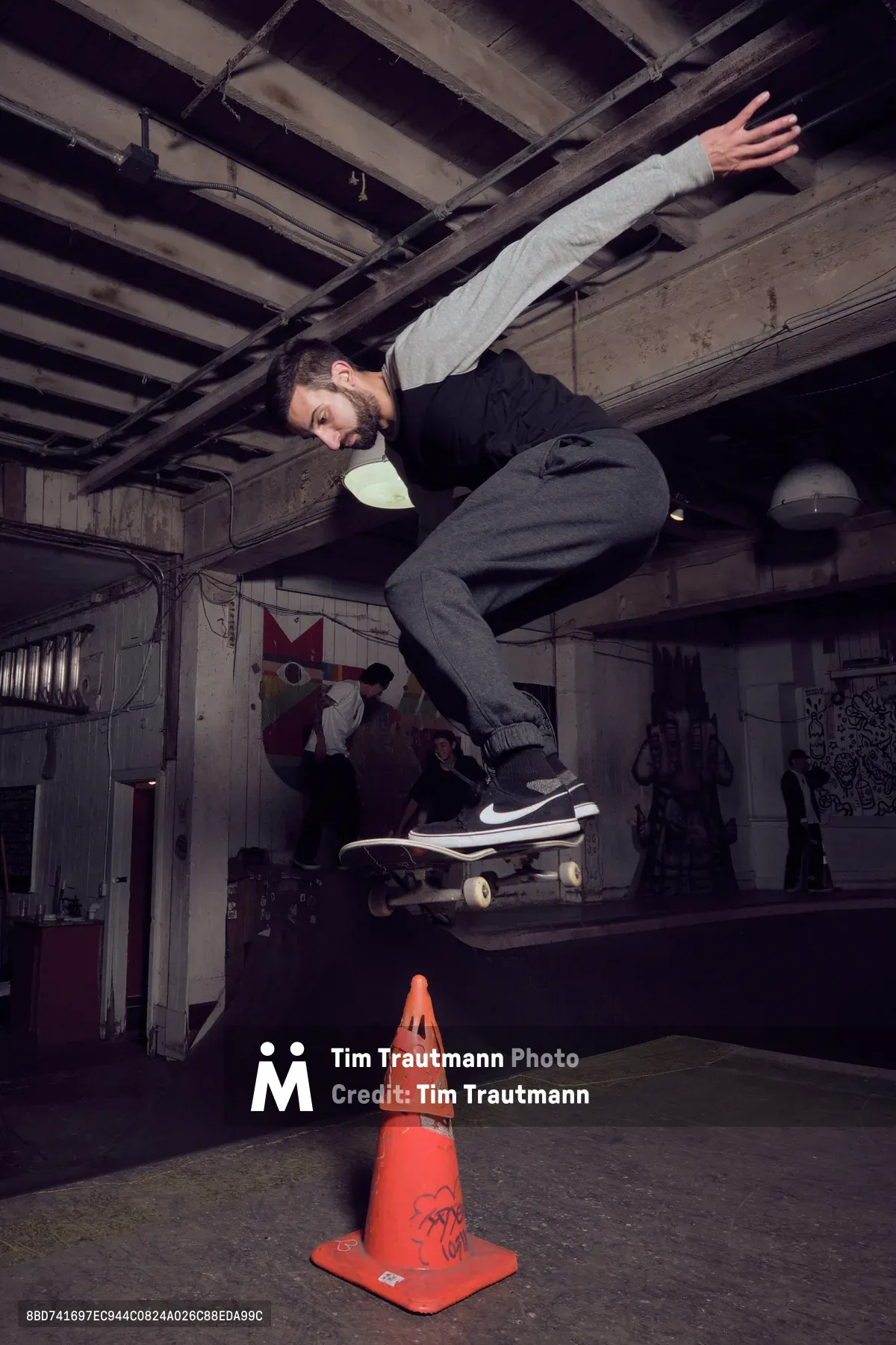 A bearded skateboarder launches into the air above an orange traffic cone in the dimly lit Commonwealth Skateboarding in Portland's Buckman neighborhood. The atmospheric industrial space features exposed wooden ceiling beams and concrete floors, creating a raw underground aesthetic. Moody lighting casts dramatic shadows while graffitied walls and other skaters in the background establish the authentic street culture setting. The frozen moment captures the skater mid-trick, his body extended with arms spread for balance as his board hovers beneath him.