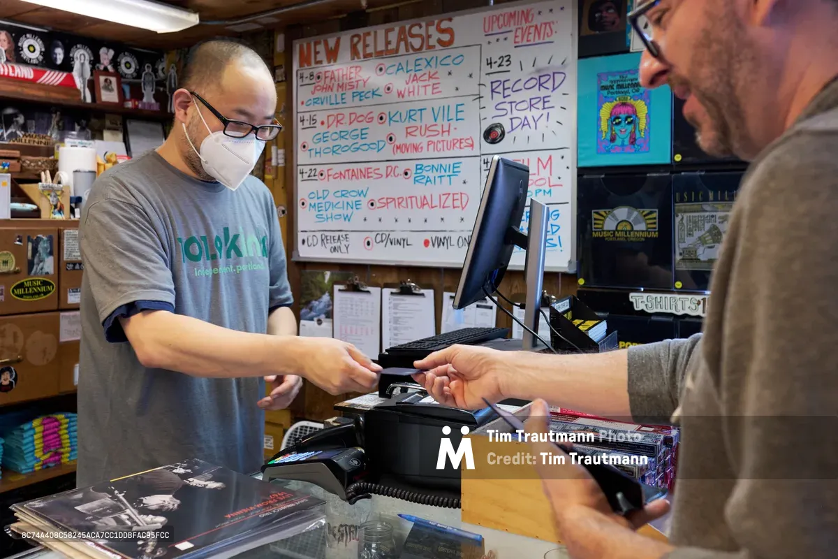 Inside Music Millennium on East Burnside during Record Store Day 2022, a masked clerk in glasses and a gray t-shirt processes a customer's purchase at the checkout counter. The intimate record store setting features handwritten new release boards advertising vinyl from artists like Dr. Dog and Kurt Vile, while shelves packed with albums and merchandise create a cozy backdrop. Warm interior lighting illuminates the exchange between the two men as physical media changes hands in this enduring bastion of music culture during the annual celebration of independent record stores.