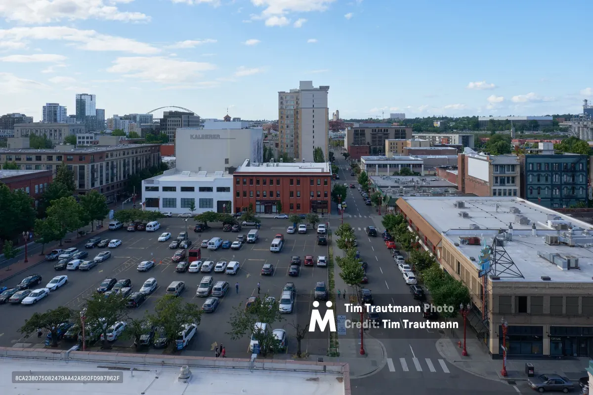 An elevated perspective captures the heart of downtown Portland's Old Town Chinatown district, where a bustling surface parking lot serves as the focal point amid a tapestry of historic brick buildings and modern structures. The late afternoon light bathes the mixed-use neighborhood in warm tones, highlighting the architectural diversity from restored industrial buildings to contemporary residential towers. Tree-lined streets radiate outward from the central plaza, while pedestrians move through the urban landscape beneath a partly cloudy Pacific Northwest sky.