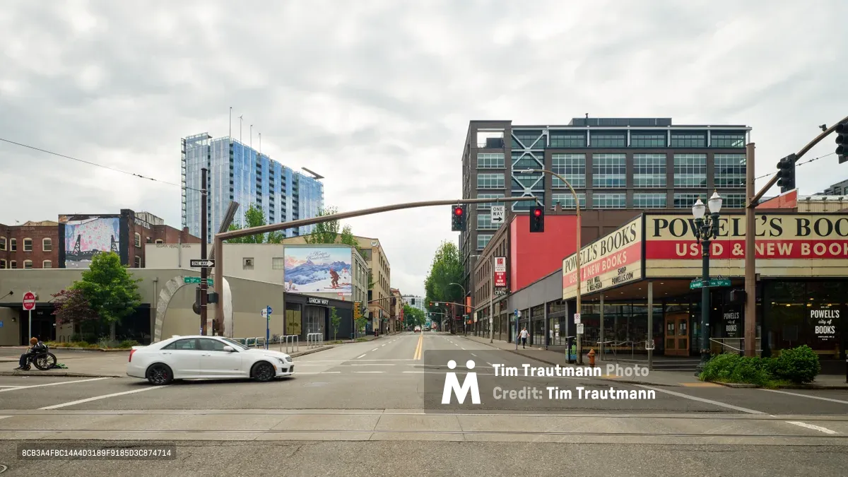 A lone white sedan crosses the eerily deserted intersection of West Burnside Street in Portland's Pearl District, capturing the profound stillness of the early COVID-19 pandemic lockdown. The iconic Powell's Books storefront dominates the right side of the frame, its familiar red and gold signage a stark contrast to the vacant sidewalks that would normally bustle with literary enthusiasts. Overcast skies mirror the somber mood of this historic moment, while modern high-rise towers loom in the background, silent witnesses to the city's sudden pause.
