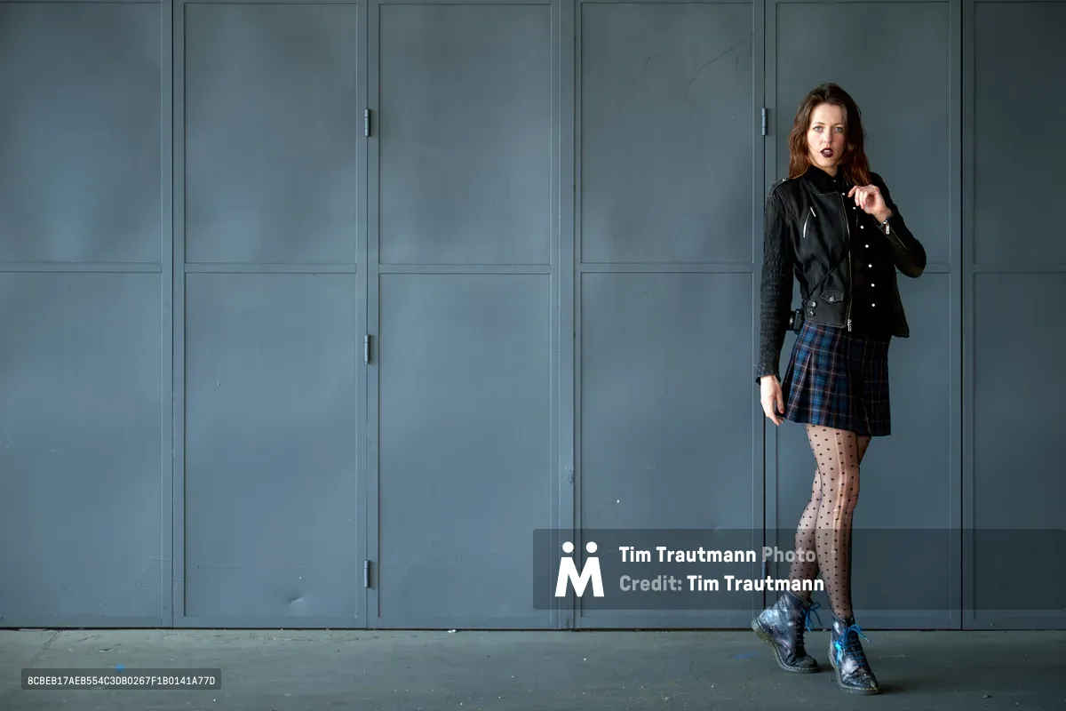 Model Rhianna Atwood strikes a contemplative pose against weathered steel lockers in Portland's industrial Central Eastside district. Her edgy ensemble of black leather jacket, plaid mini skirt, and polka-dot tights creates a striking contrast against the utilitarian blue-gray backdrop. The natural lighting casts subtle shadows, emphasizing the juxtaposition between punk-inspired fashion and the stark industrial architecture of Southeast Portland's warehouse district.