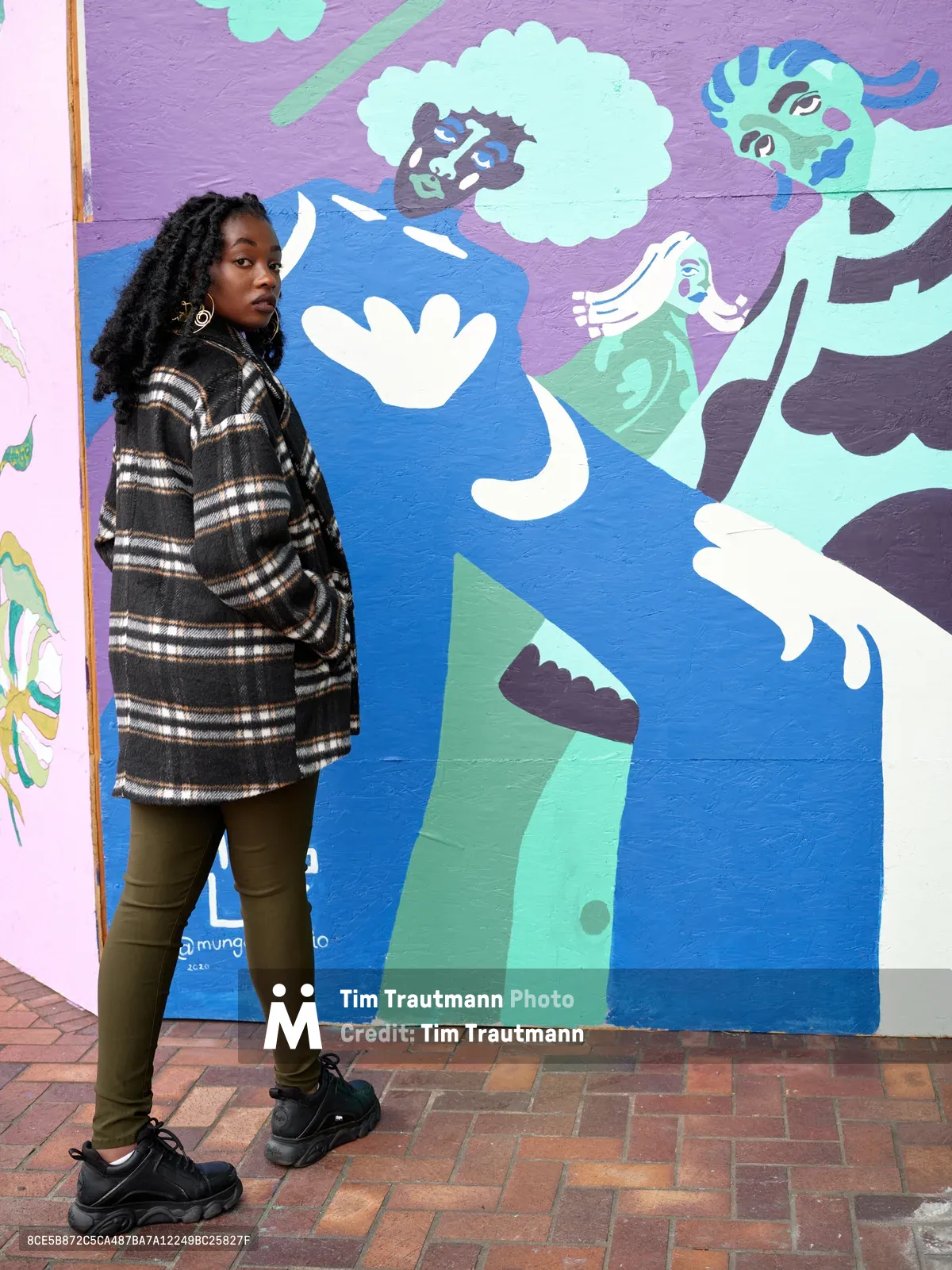 Naomi Likayi stands confidently beside her colorful community mural painted on the boarded Portland World Trade Center, commissioned by the Portland Street Art Alliance. The artist wears a plaid coat and locs, positioned against the whimsical artwork featuring abstract figures in blues, purples, and greens that transform the urban wall into a canvas of hope. Her direct gaze and relaxed stance create an intimate portrait that captures both the artist and her public art contribution to Portland's cultural landscape.