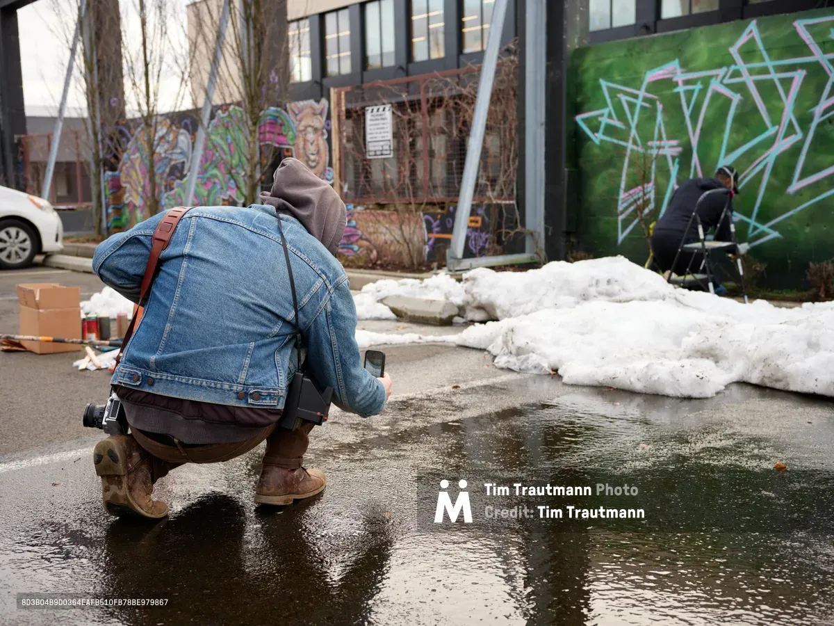 A photographer crouches low to capture a street muralist at work in the parking lot of Clay Creative in Southeast Portland's Hosford-Abernethy neighborhood. The scene unfolds against a backdrop of vibrant graffiti-covered walls, with melting snow creating dark puddles that mirror the overcast winter sky. The intimate composition draws viewers into the creative process, highlighting the dedication required to transform urban spaces into canvases of expression.
