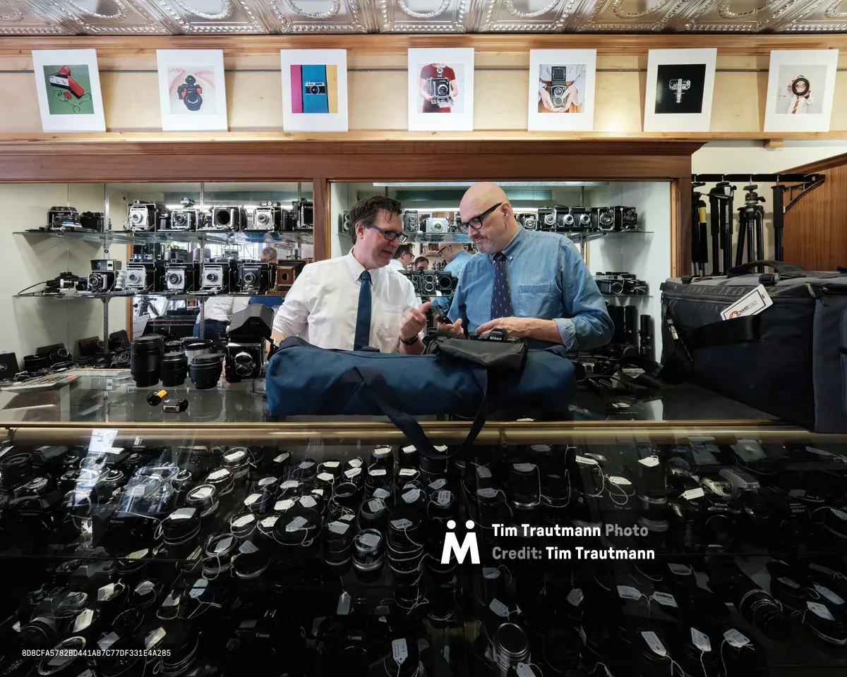 Mike Knight and Arthur Ruckle examine vintage cameras amid the organized chaos of Blue Moon Camera & Machine's Portland storefront. Warm tungsten light bathes the ornate tin ceiling and illuminates rows of meticulously arranged vintage cameras displayed on glass shelves, while hundreds of lenses carpet the floor below. The two specialists lean over their inspection table in focused collaboration, surrounded by decades of photographic history in this legendary Pearl District camera shop.