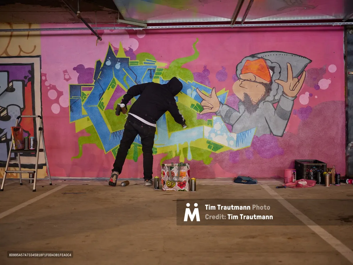 A muralist in black clothing works intently on a vibrant wall piece within the industrial confines of Clay Creative's parking garage in Southeast Portland. The dynamic composition features bold geometric forms in electric blues, acid greens, and hot pinks against the raw concrete backdrop, while spray paint cans and art supplies scatter across the floor. Exposed ceiling infrastructure and fluorescent lighting create an authentic underground studio atmosphere, capturing the transformation of utilitarian space into creative canvas.