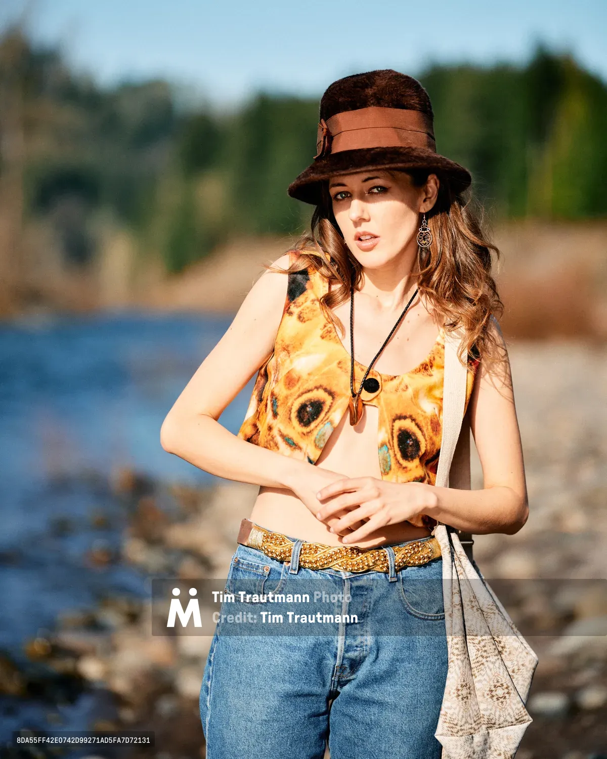 A young woman poses confidently along Oregon's Floodplain Trail, wearing a distinctive butterfly-print vest over a cream top, complemented by high-waisted denim and a russet felt hat with leather band. The warm afternoon light illuminates her flowing brown hair and ornate earrings, while the Columbia River's blue waters and lush Multnomah County landscape create a dreamy, out-of-focus backdrop. Her relaxed stance and gentle expression embody the free-spirited essence of Pacific Northwest outdoor culture.