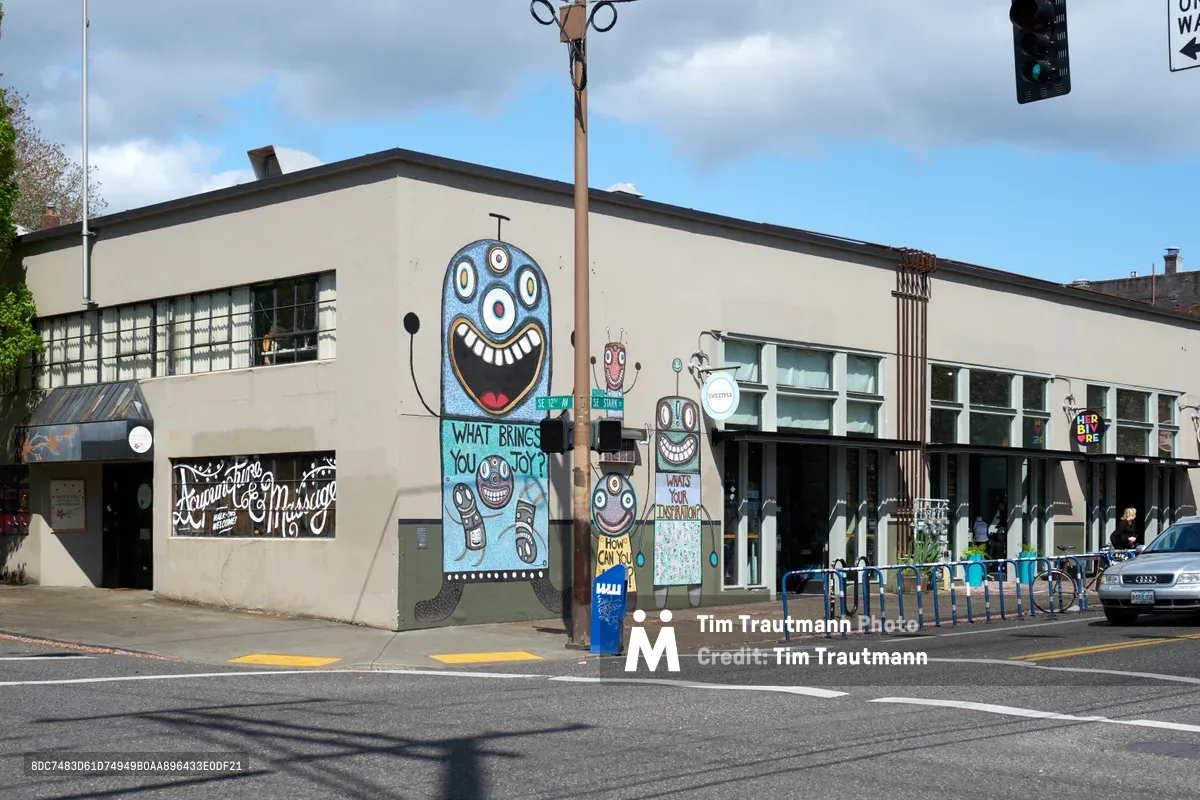 A vibrant mural transforms the corner building at SE Stark Street and SE 12th Avenue in Portland's Central Eastside district. The playful artwork features a large blue cartoon face asking "What brings you joy?" alongside smaller character illustrations, creating an engaging dialogue with passersby. Overcast skies provide soft, even lighting that enhances the mural's bold colors against the neutral stucco facade, while urban infrastructure elements like traffic lights and bike racks frame the scene.