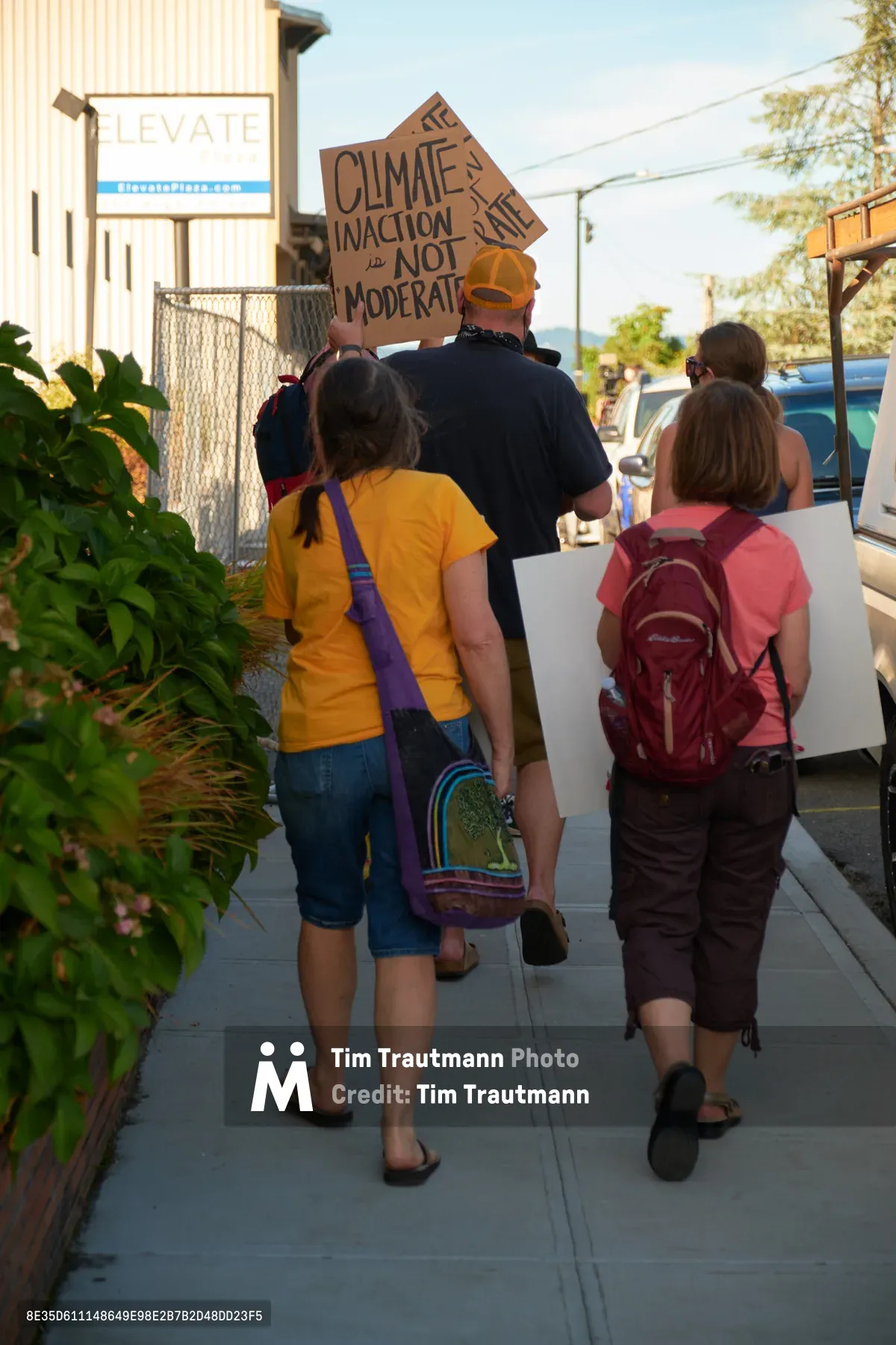 A small group of environmental protesters walks along McLoughlin Promenade in Oregon City, carrying handmade signs advocating for climate action. The late afternoon sun casts long shadows across the concrete sidewalk as the demonstrators make their way past local businesses, including a visible LEVATE sign on a commercial building. The informal procession captures the grassroots nature of climate activism in small-town Oregon, with participants of varying ages united in their call for environmental urgency.