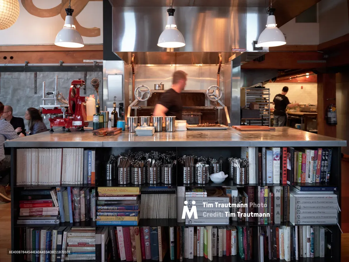A striking open kitchen design combines culinary workspace with literary collection in this Portland restaurant. Professional chefs move purposefully behind a gleaming stainless steel island, while hundreds of cookbooks line integrated shelving below the counter. Industrial pendant lights cast warm illumination over the bustling workspace, where diners can observe the choreographed dance of professional food preparation amid towering volumes of culinary knowledge.