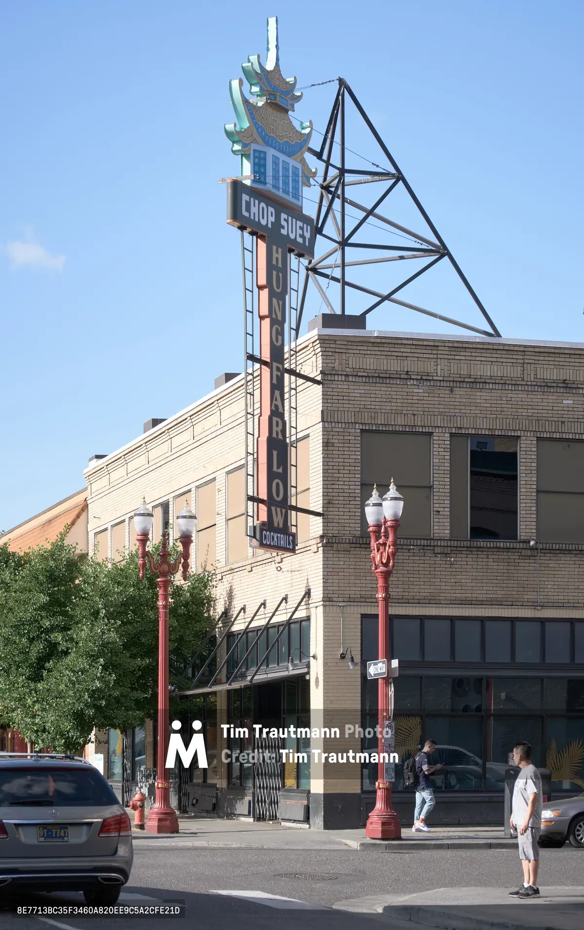 A towering vintage neon sign for the Hung Far Low restaurant dominates the streetscape in Portland's historic Chinatown district. The elaborate sign features traditional Chinese pagoda motifs and colorful neon tubing against a clear blue sky. Below, pedestrians walk along the sidewalk past the brick commercial buildings that define this culturally significant neighborhood, while period street lamps and parked cars complete the urban scene.