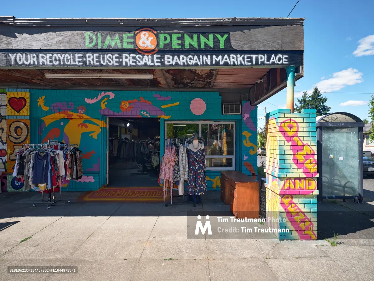 A vibrant thrift shop facade on Northeast 15th and Killingsworth in Portland's Alberta neighborhood bursts with kaleidoscopic murals across weathered walls. Clothing racks spill onto the sidewalk beneath hand-painted signage declaring this a recycle-reuse-resale bargain marketplace. The turquoise, orange, and pink street art transforms the humble storefront into a celebration of sustainable shopping culture.