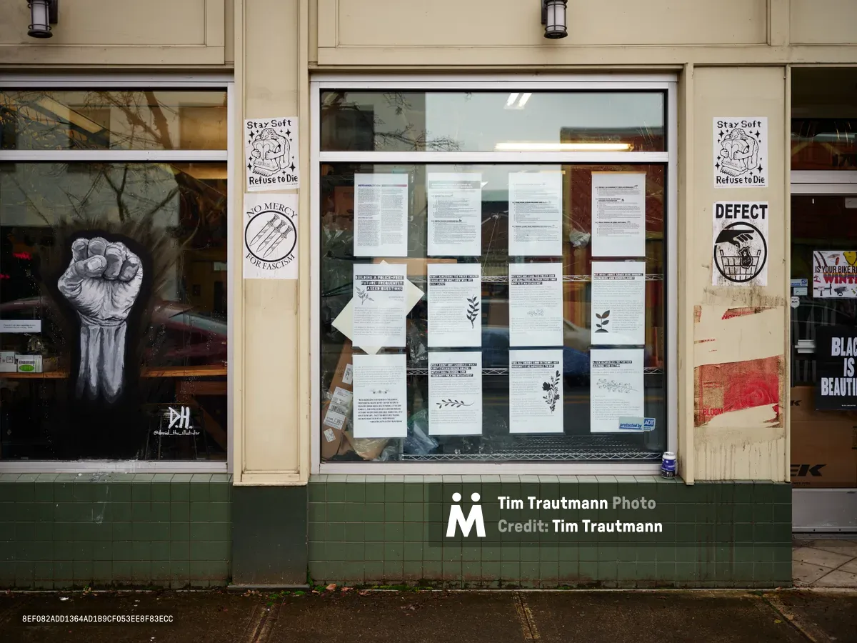 A weathered storefront window displays a powerful collection of antifascist posters and political documents along Northeast Alberta Street in Portland's Vernon neighborhood. The cream-colored facade frames multiple protest flyers, including a striking raised fist illustration and "Stay Soft Refuse to Die" messaging, while "NO MERCY FOR FASCISTS" and "DEFECT" posters anchor the visual narrative. Green subway tiles at the building's base contrast with the urgent white papers pressed against glass, creating a raw street-level gallery of grassroots activism.