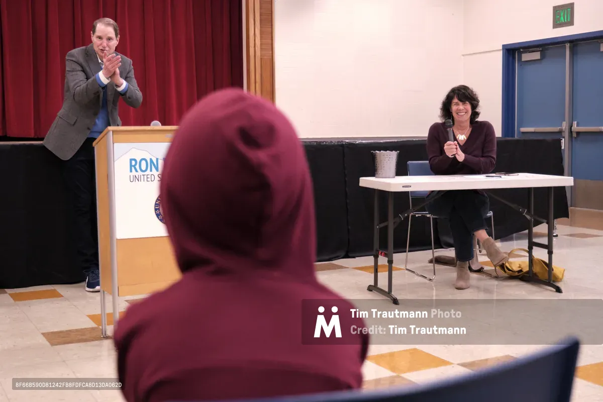 Senator Ron Wyden gestures expressively from behind a podium bearing his name, engaging with constituents during a town hall meeting at Robert Gray Middle School in Portland's Hillsdale neighborhood. The intimate setting features burgundy curtains and polished floors, with a moderator seated at a white table nearby. A community member in a deep red hooded sweatshirt sits in the foreground, representing the engaged local audience participating in this democratic dialogue.