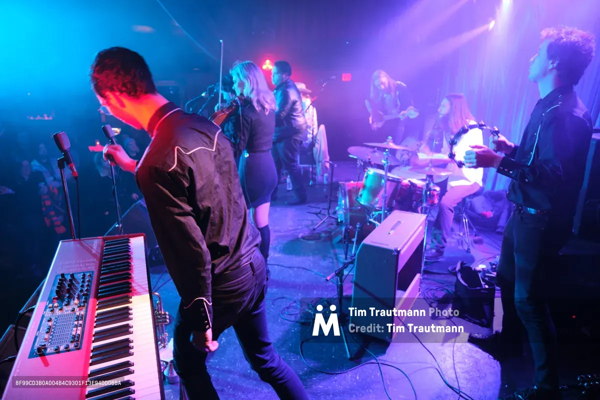 Roselit Bone commands the cramped stage at Portland's legendary Dante's venue, their silhouettes carved by piercing purple and cyan stage lights that slice through the smoky darkness. The band's keyboardist leans into his vintage synthesizer while the guitarist and drummer create a wall of sound behind him, all bathed in the venue's signature neon glow. The intimate basement setting amplifies the electric energy, with cables snaking across the worn floor and amplifiers crowding the small performance space.